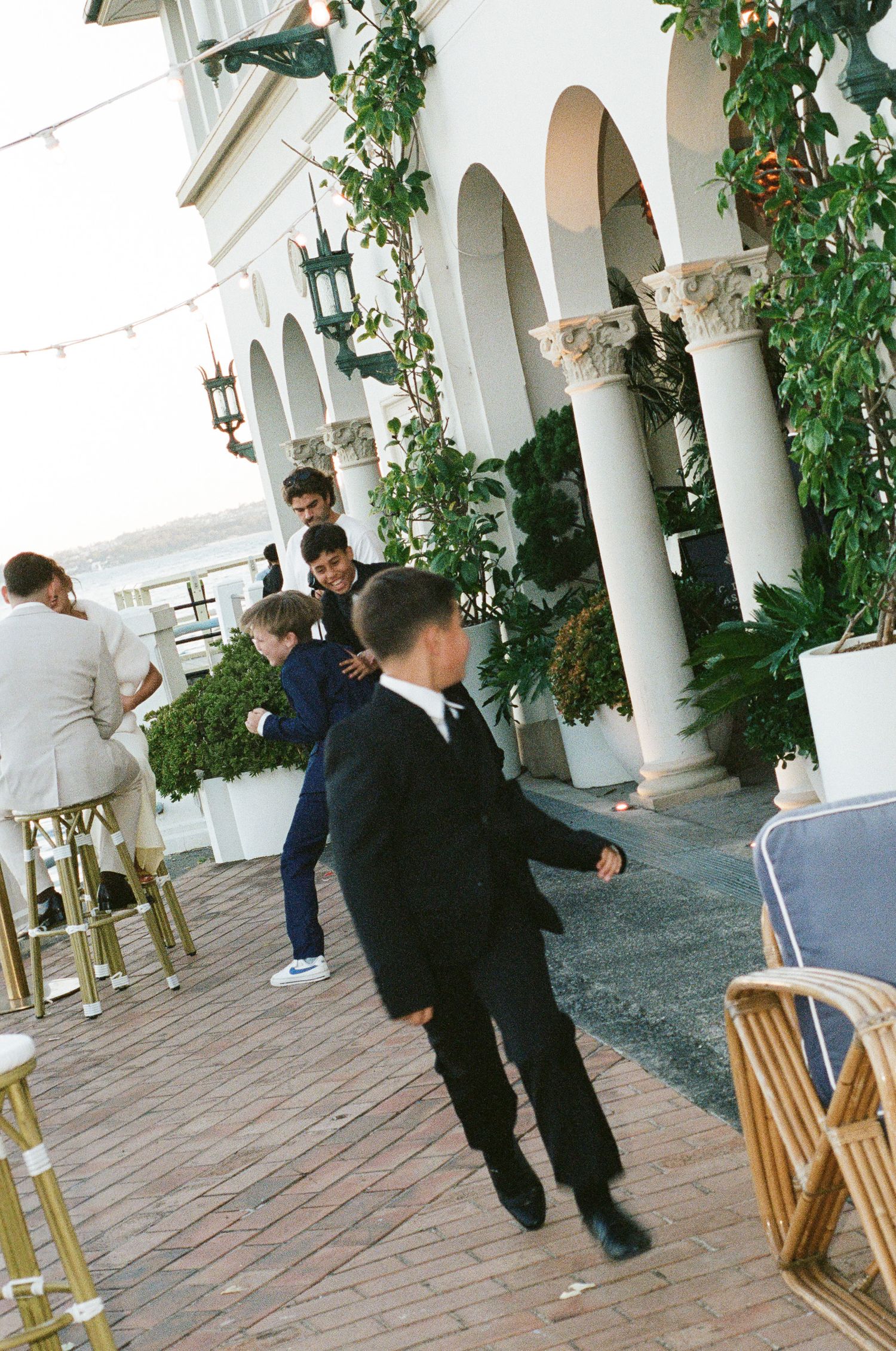 Boys in dark suits play on a brick terrace beside white arched columns, climbing greenery, and rattan chairs at an outdoor event.