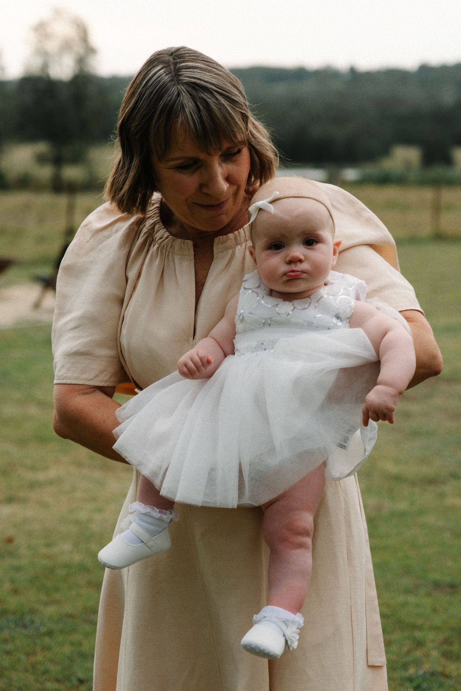 A baby wearing a white tulle dress and socks is held outdoors by someone in a cream-colored dress.