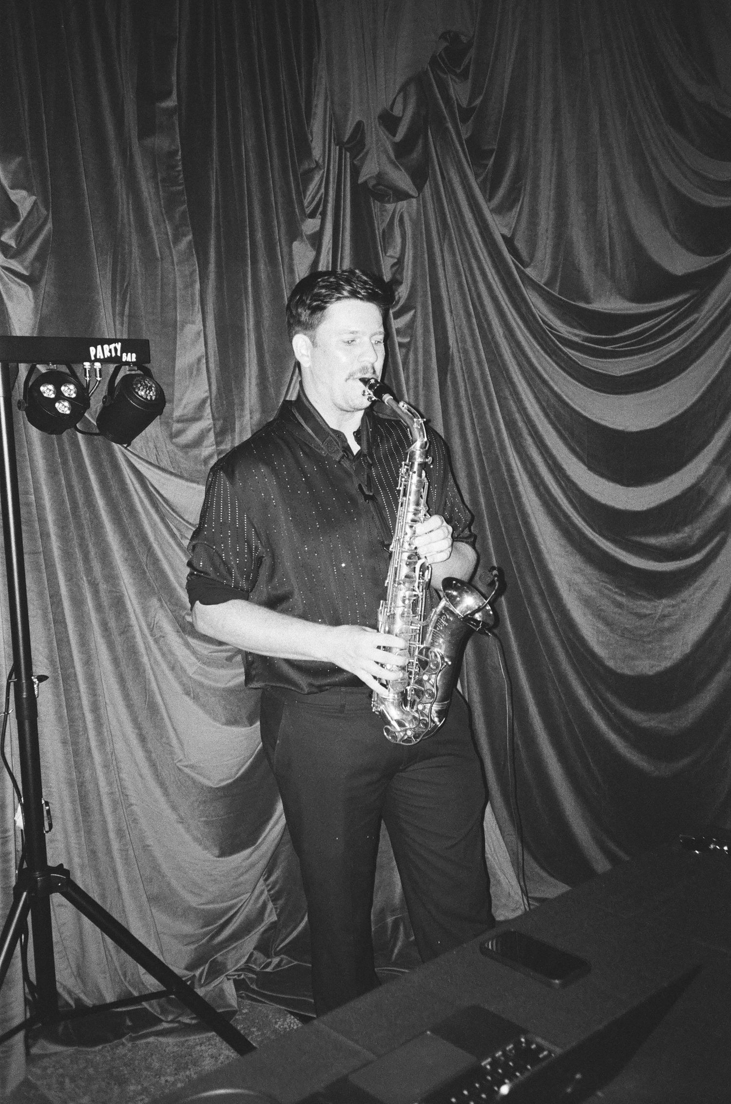 A musician performs with a saxophone on stage against draped curtains in a black and white photograph.
