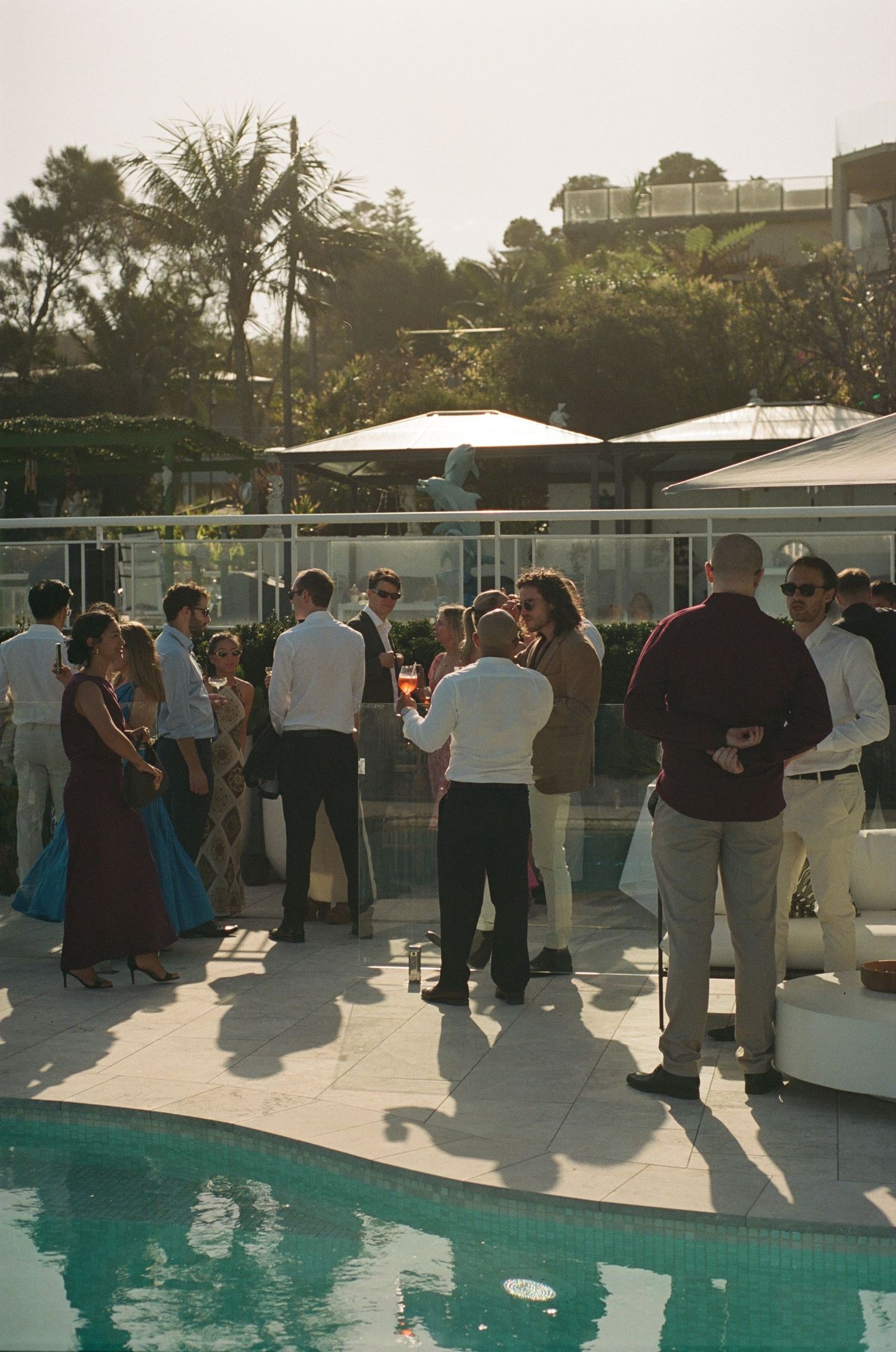 People gathered at an outdoor poolside party during sunset with umbrellas and mingling guests.
