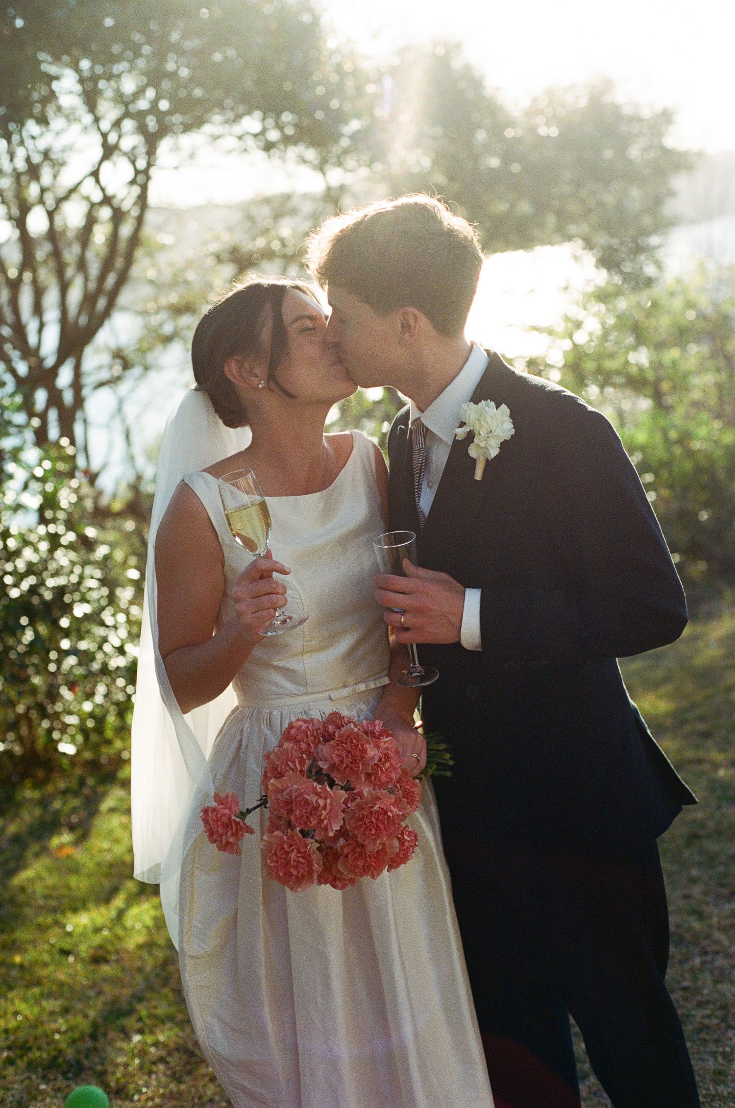 Bride in a white dress and veil kisses groom in a navy suit while holding champagne and pink carnations in a sunlit garden.