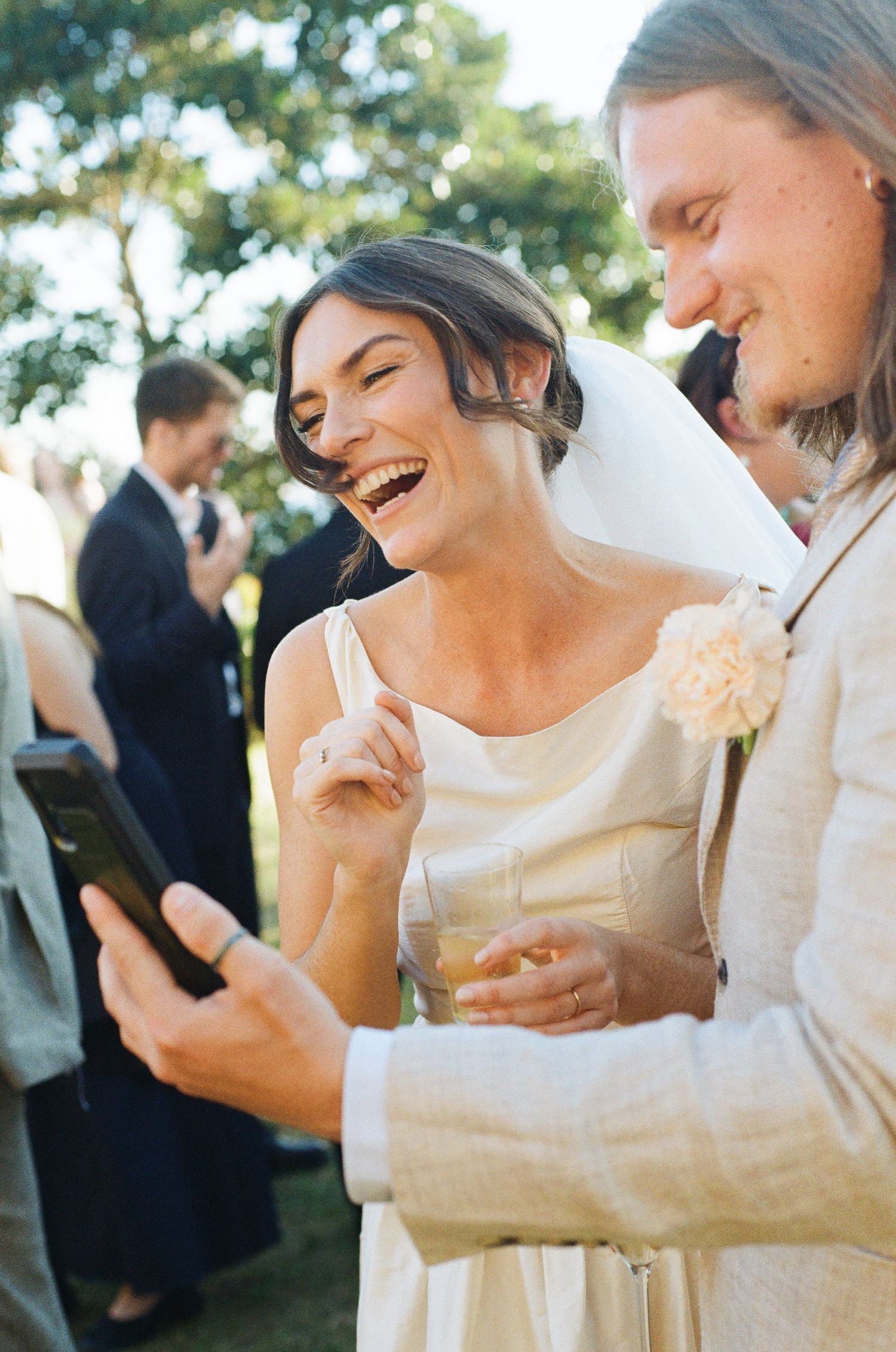 A laughing bride in a white dress and veil holds a drink while a man with a peach boutonniere shows her his phone at an outdoor wedding.