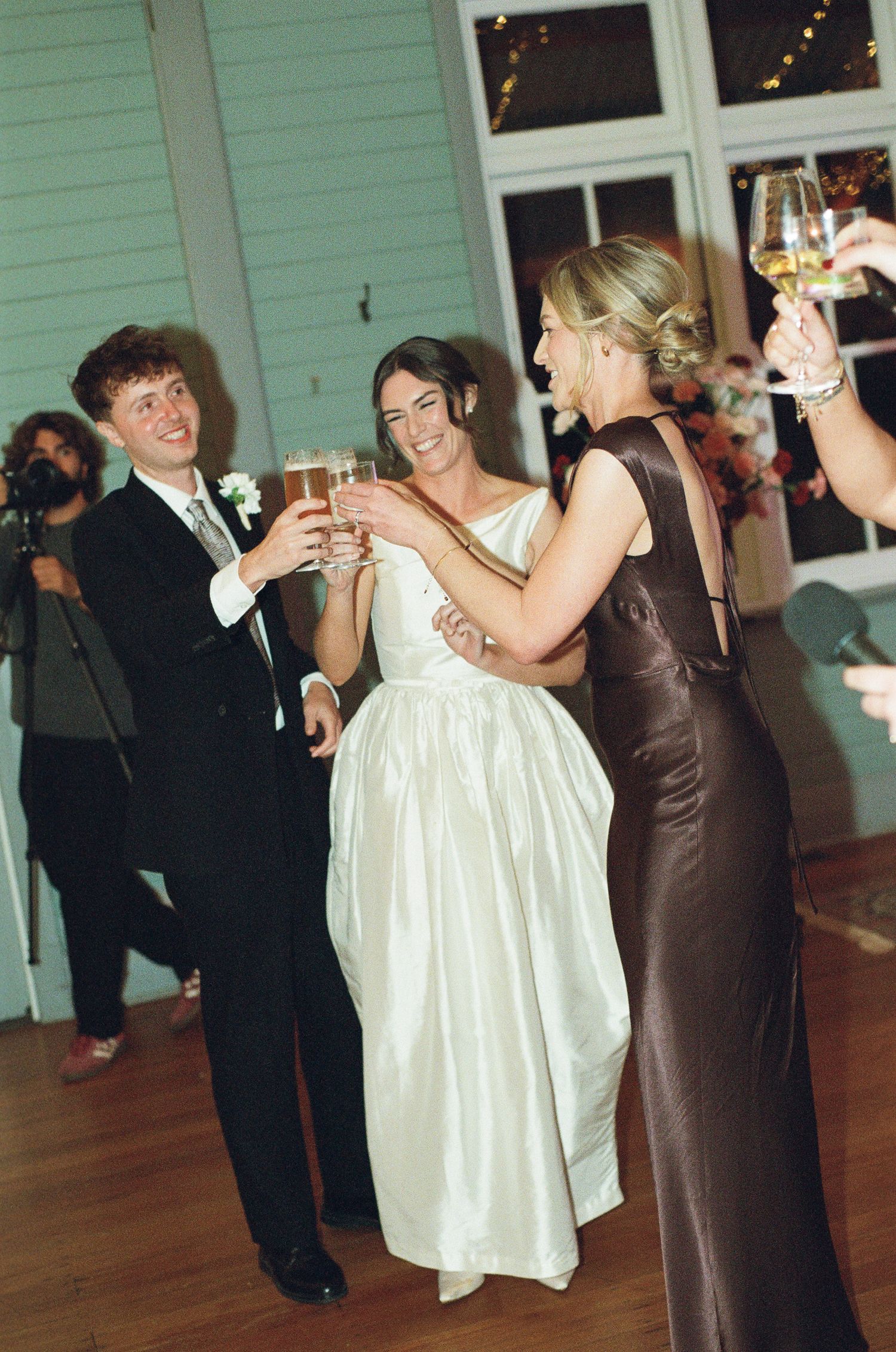 Bride in a white gown and groom in a dark suit raise champagne glasses in a toast with a woman in a brown dress at a wedding reception.