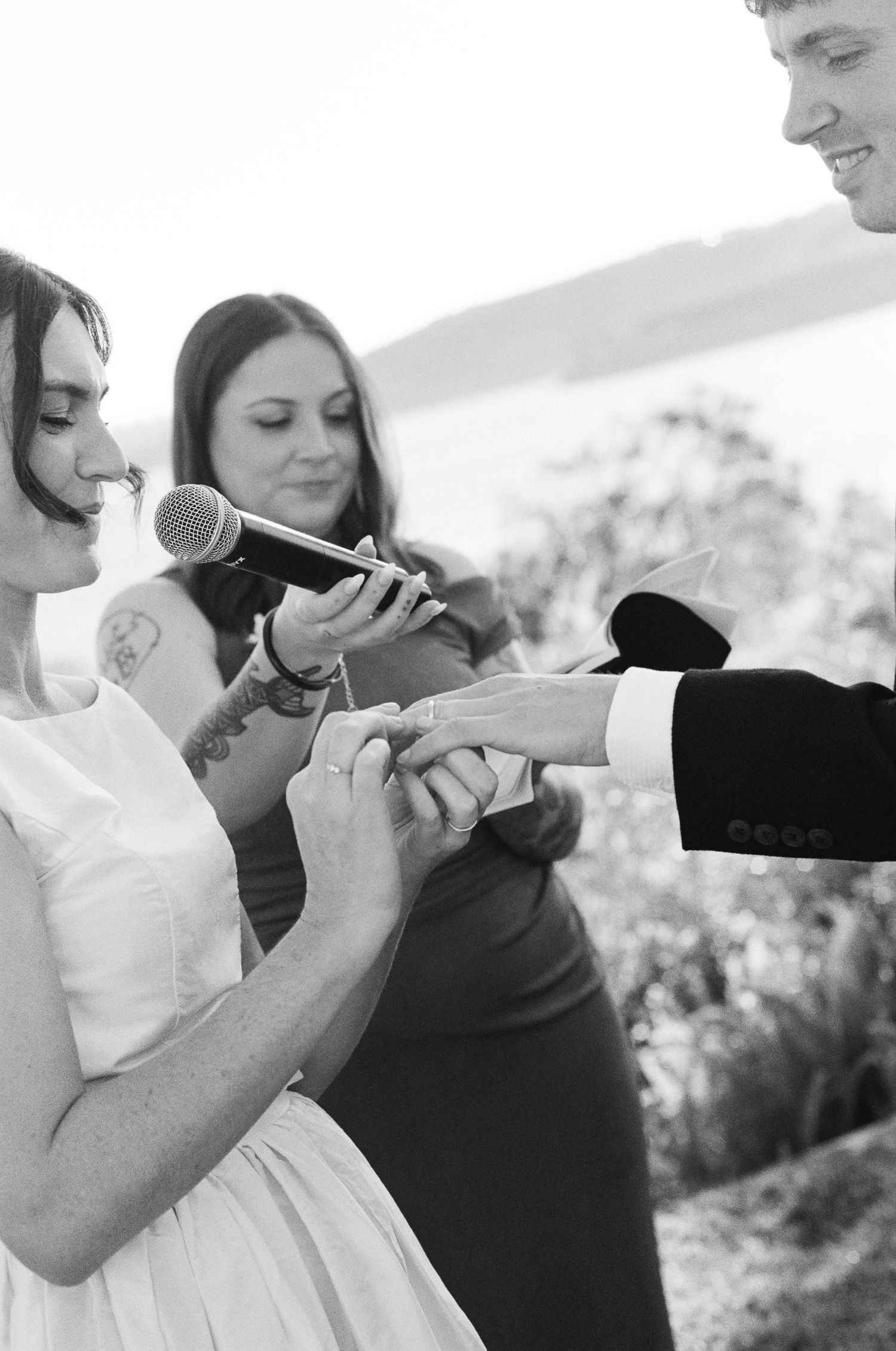 Black and white photo of a bride and groom exchanging rings during an outdoor wedding ceremony as the officiant holds a microphone.