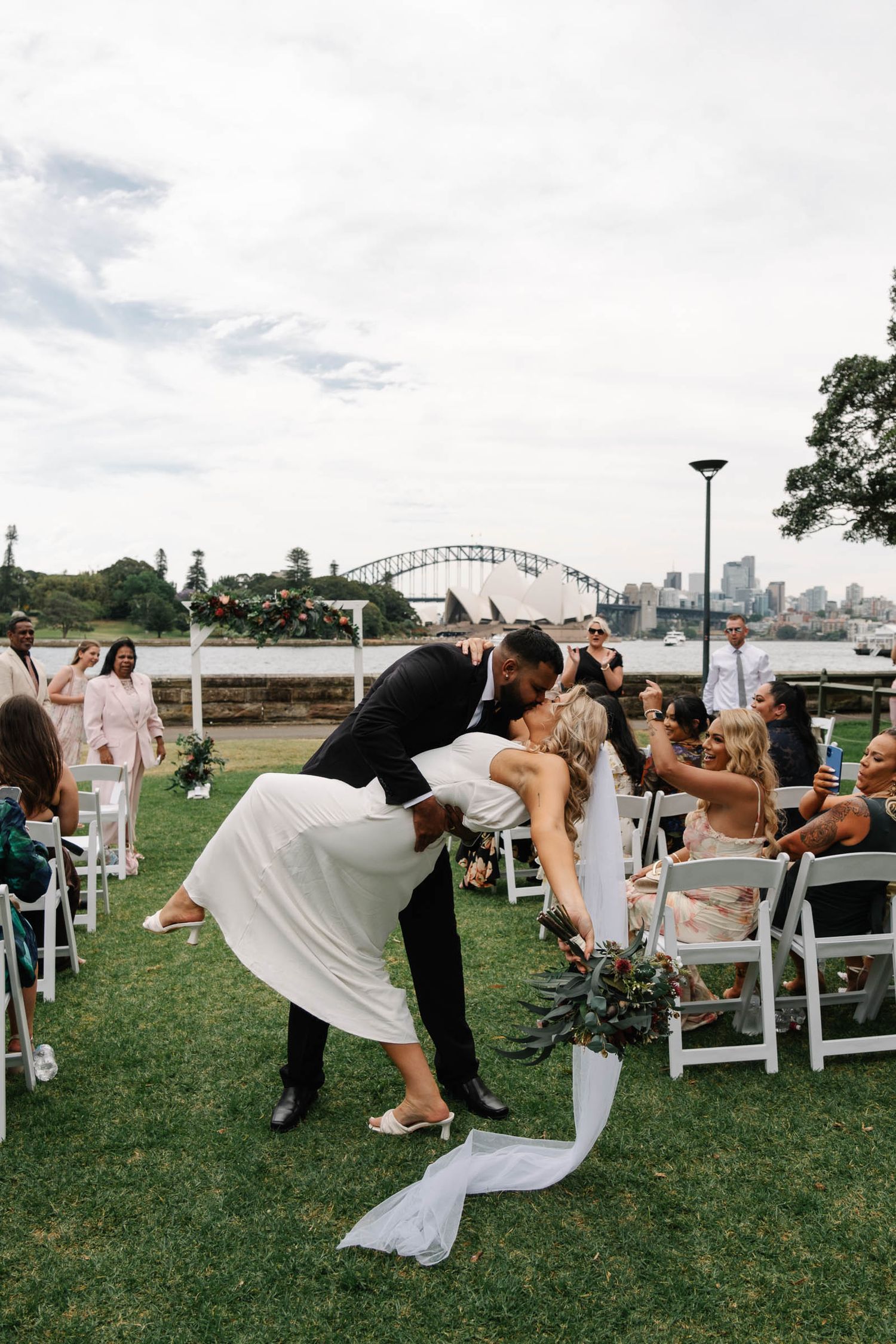 Wedding guests dip and dance together on a grassy lawn with the Sydney Harbour Bridge visible in the background.