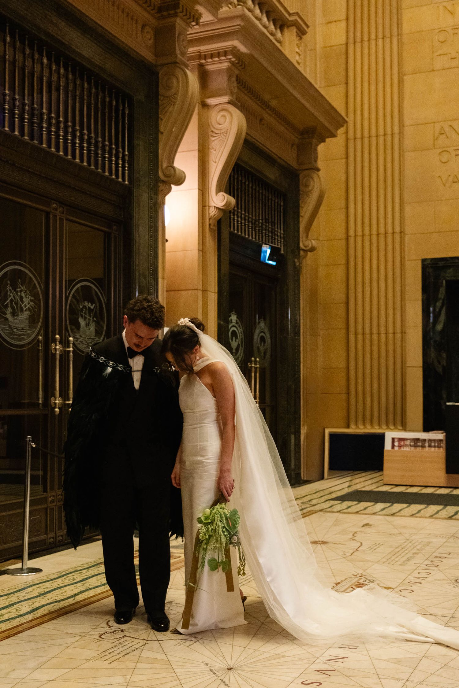 A wedding couple stands in an elegant hallway with ornate columns and marble floors, the bride's long veil trailing behind.