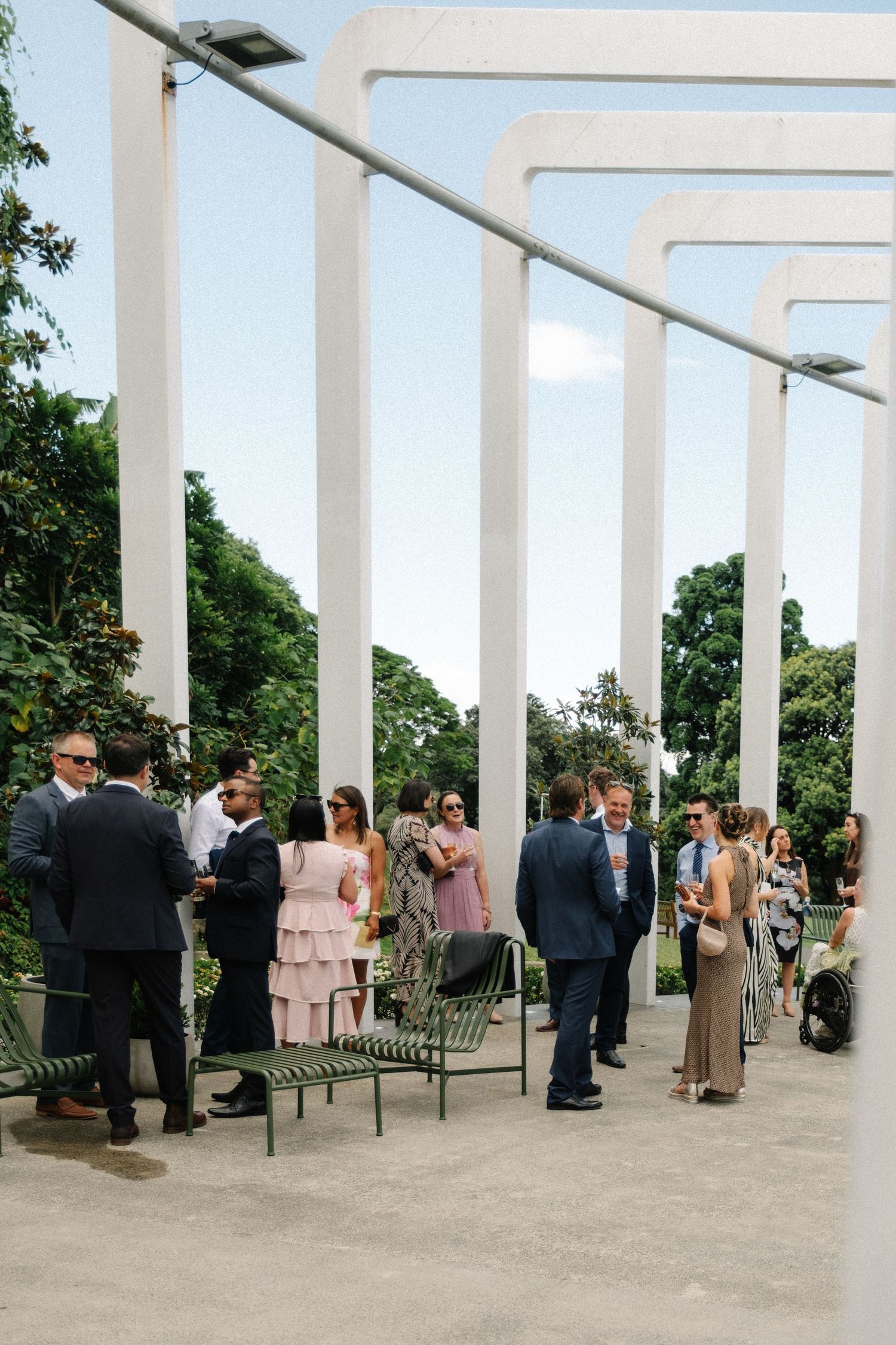 Wedding guests gather under a modern white pergola structure during an outdoor reception ceremony.