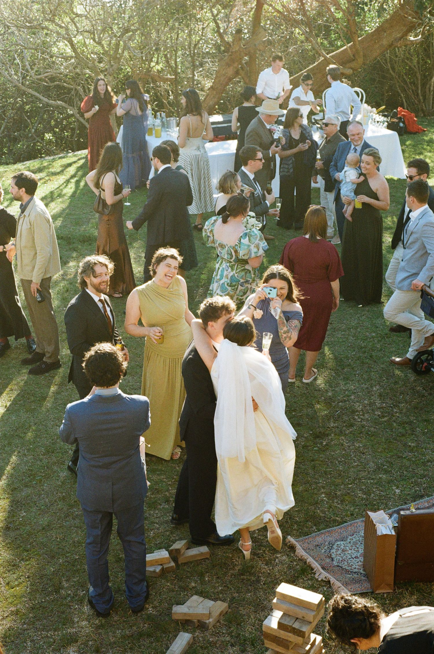 Outdoor wedding reception on green lawn with guests mingling, bride and groom sharing a kiss in the center.