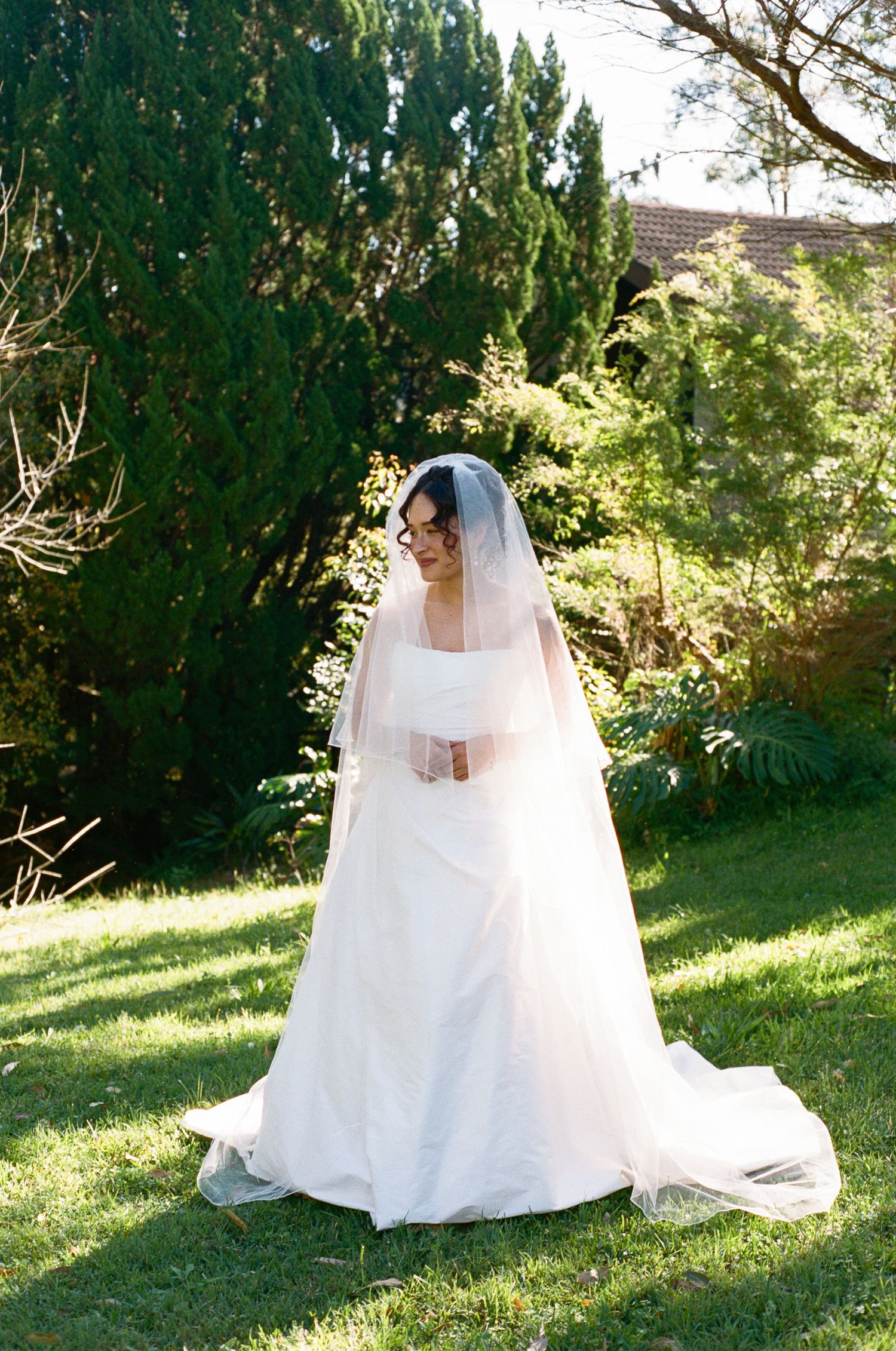 Bride in a flowing white gown and long sheer veil stands on sunlit grass surrounded by lush green trees and garden foliage.