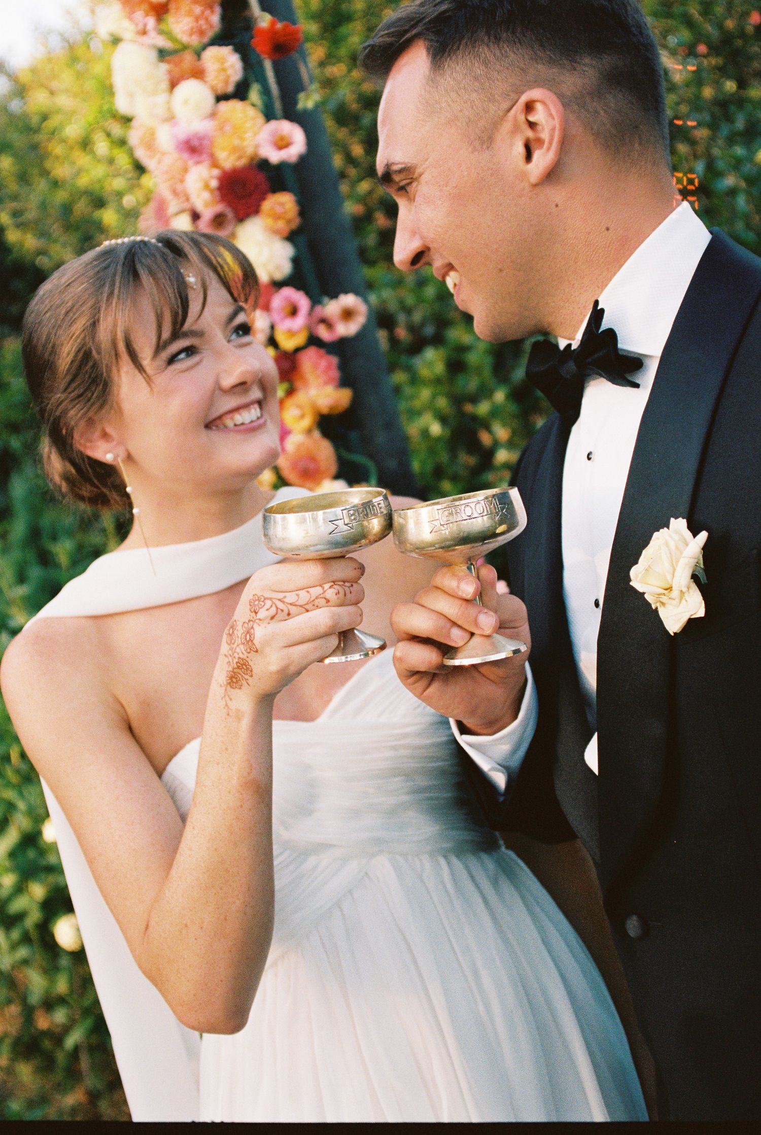 A smiling bride in a white strapless gown and a groom in a black tuxedo toast with silver goblets in a garden with colorful flowers.