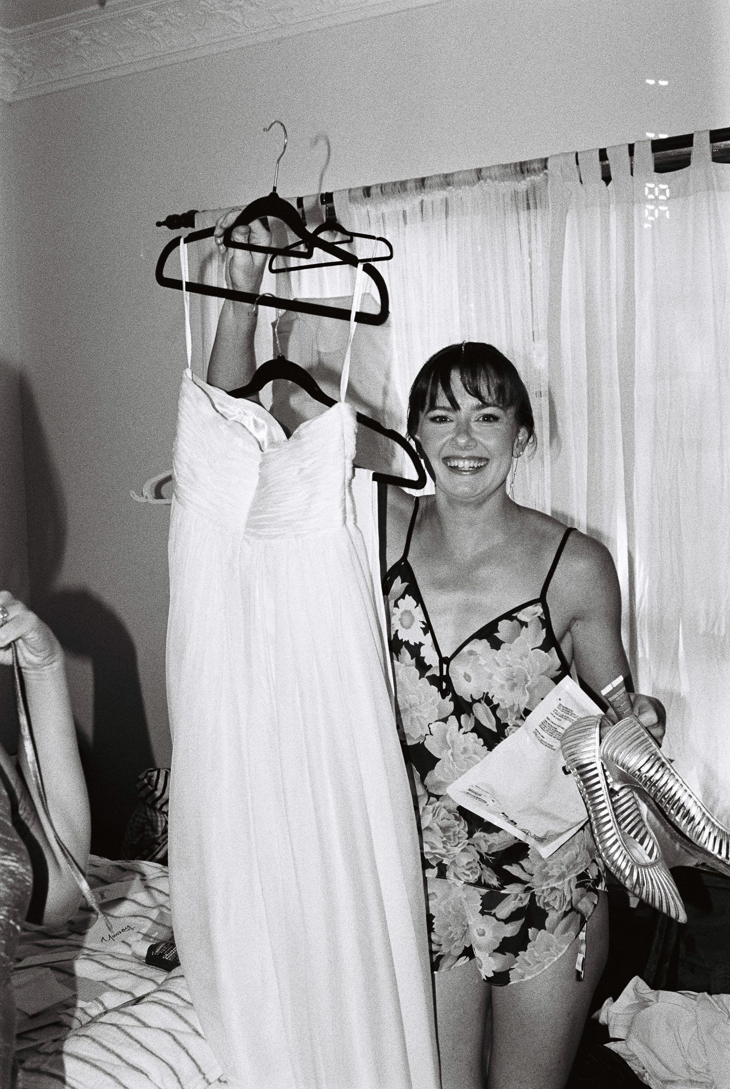 A smiling woman in a floral slip holds up a white wedding dress on a hanger and a pair of silver heels in a bright room.