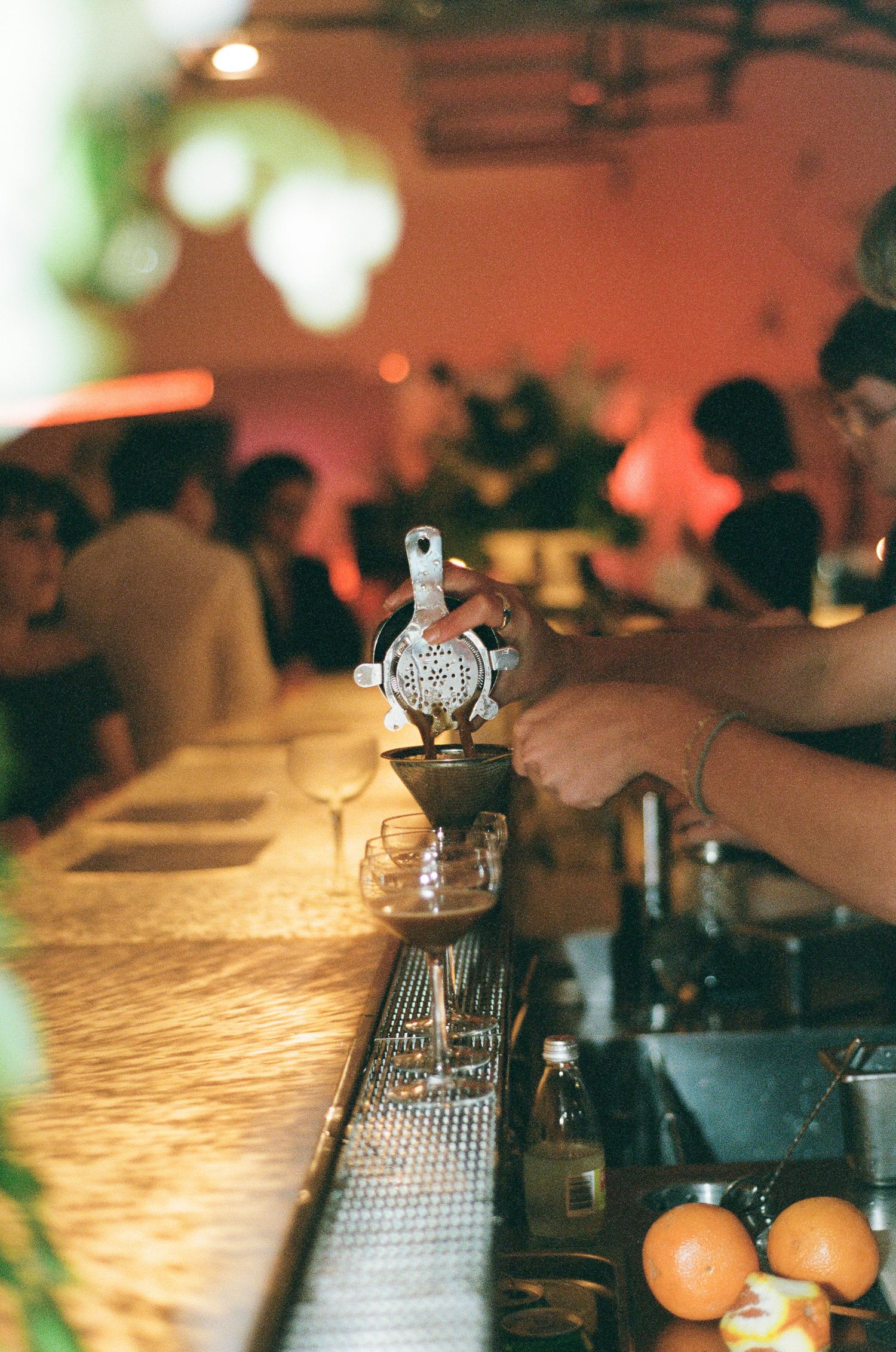 Bartender strains a cocktail through a hawthorne strainer into a coupe glass at a warmly lit bar with guests and fresh oranges nearby.