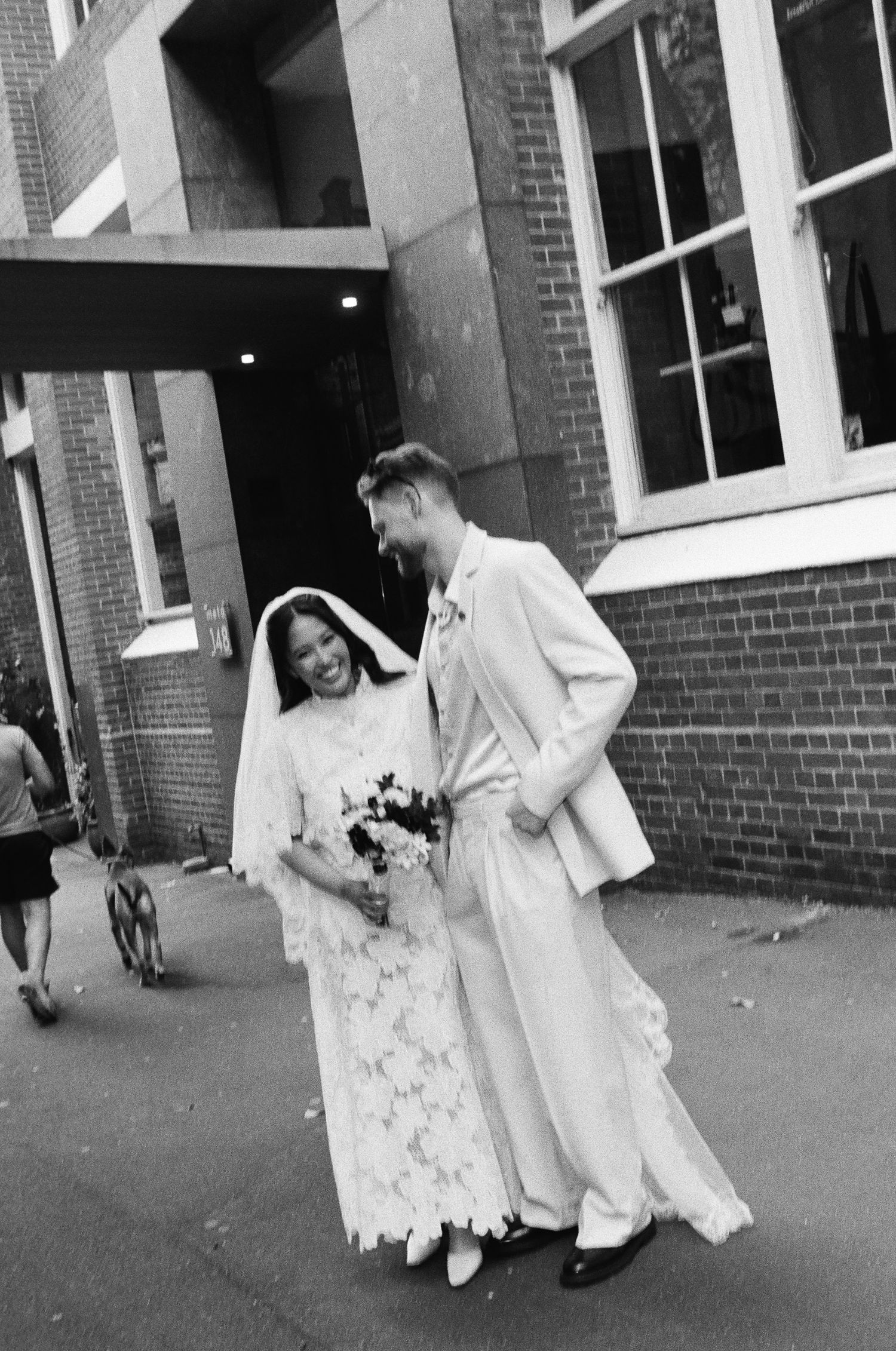 Black and white photo of a laughing bride in a lace gown and veil walking arm in arm with a groom in a light suit outside a brick building.