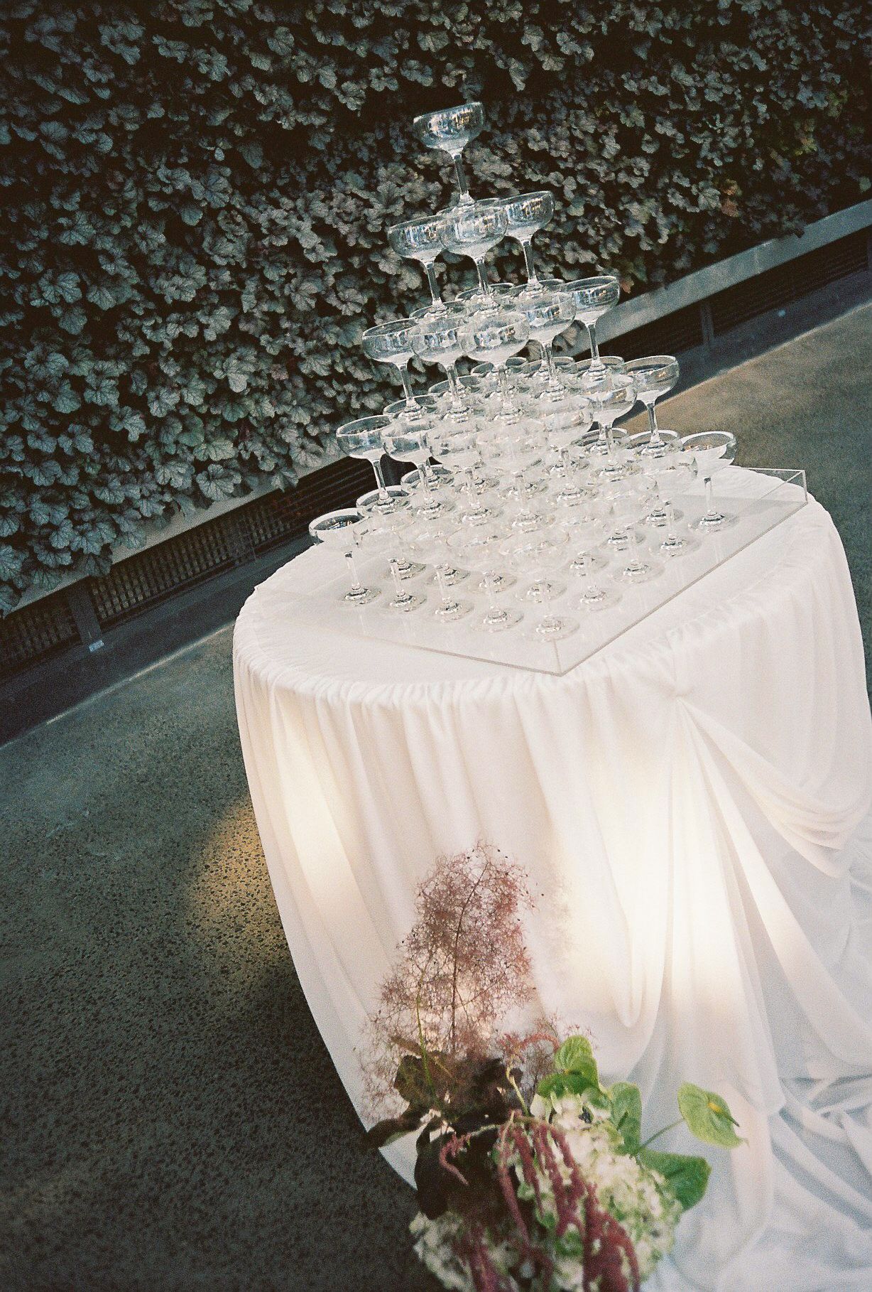 Champagne glasses arranged in a pyramid stack on white tablecloth with soft lighting and floral decorations.