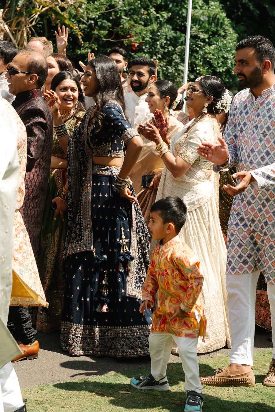 A festive outdoor gathering shows people in traditional Indian attire celebrating and dancing on a sunny day.
