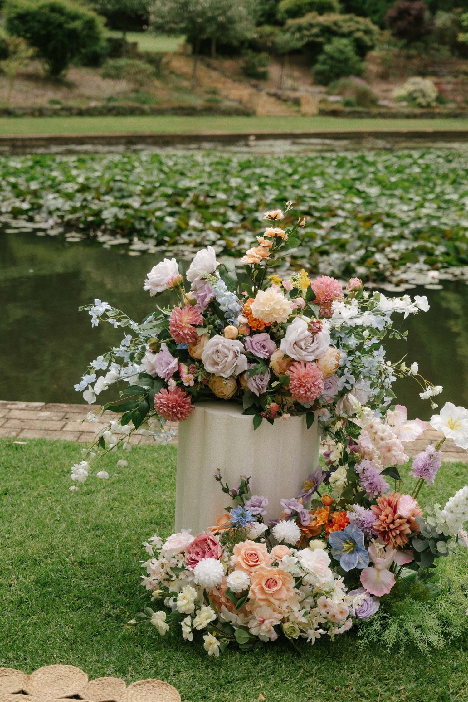 Elegant floral arrangements with pink and white blooms create a romantic garden display around a serene pond setting.