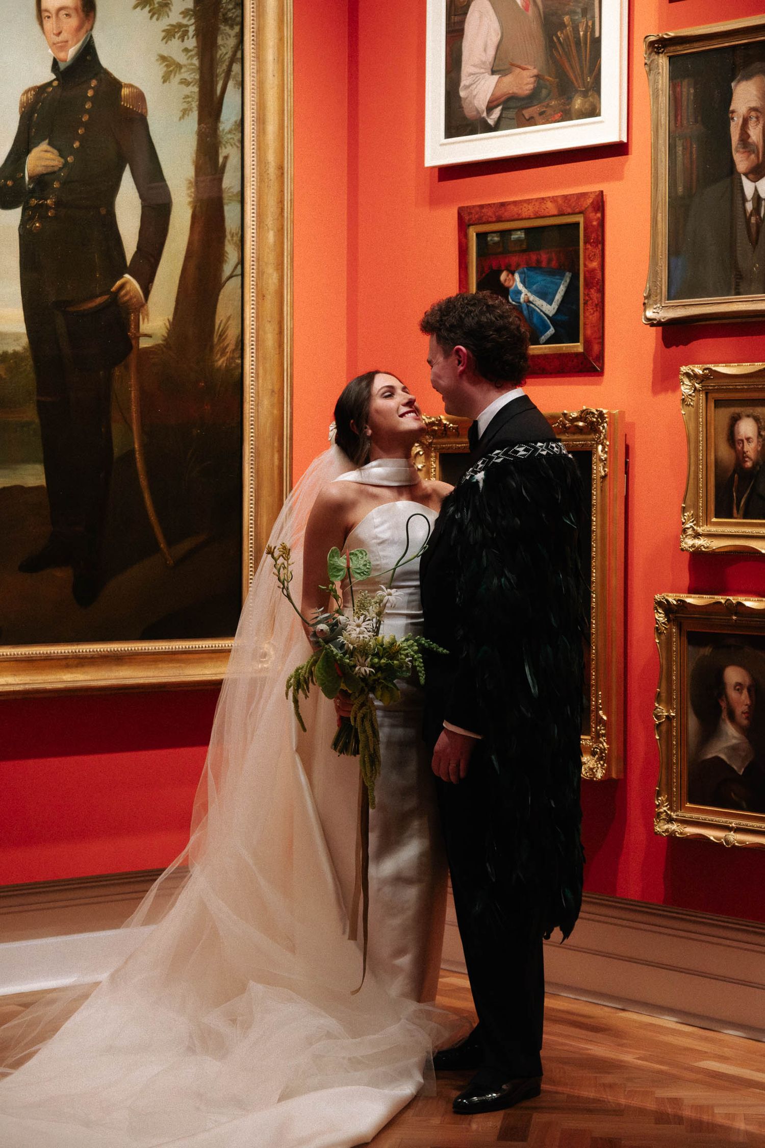 A wedding couple dances in an elegant art gallery with red walls and historic portrait paintings.