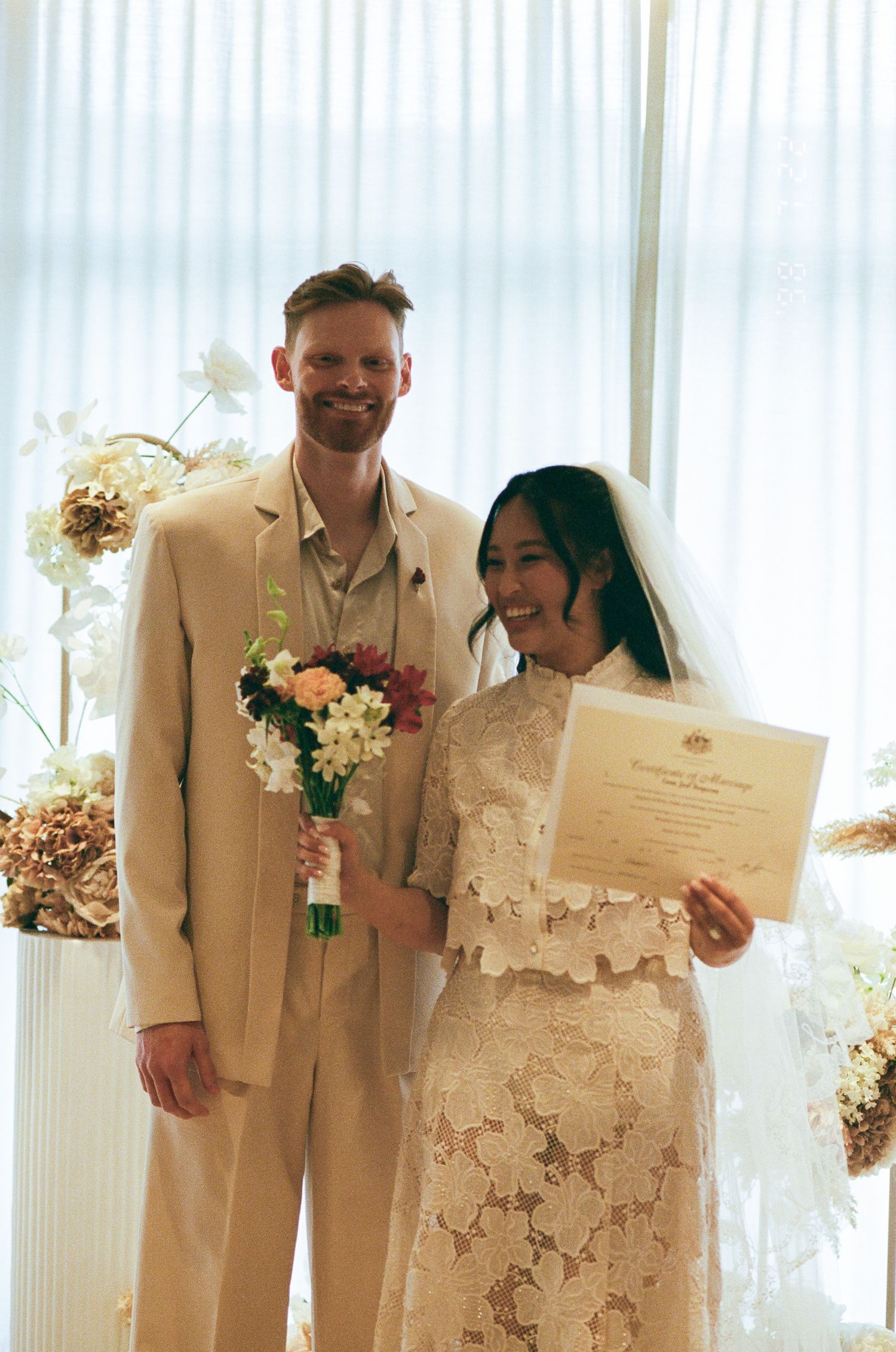 Smiling newlyweds in a beige suit and white lace dress with veil hold a bouquet and marriage certificate beside floral decor.