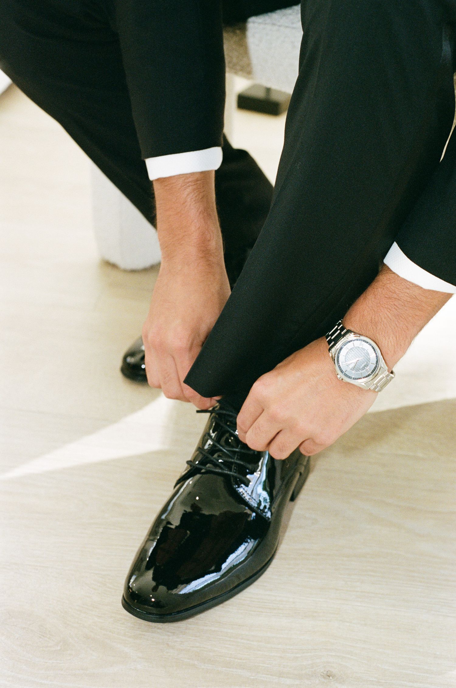 Groom in black tuxedo trousers and a silver wristwatch ties the laces of his polished black patent leather dress shoes.