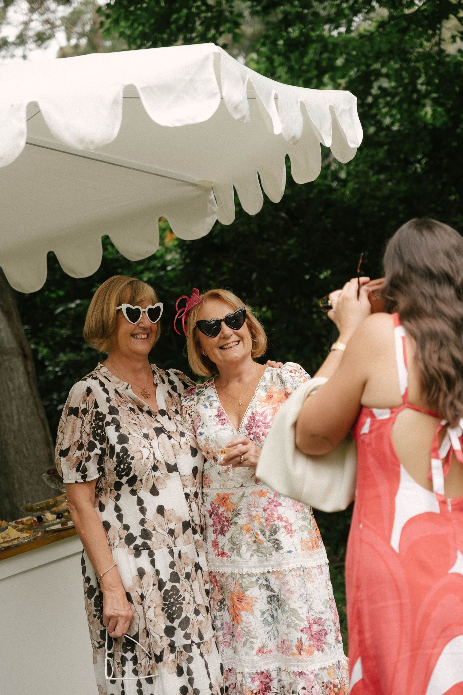 Three women in summer dresses and sunglasses laugh together under a white umbrella at an outdoor event.