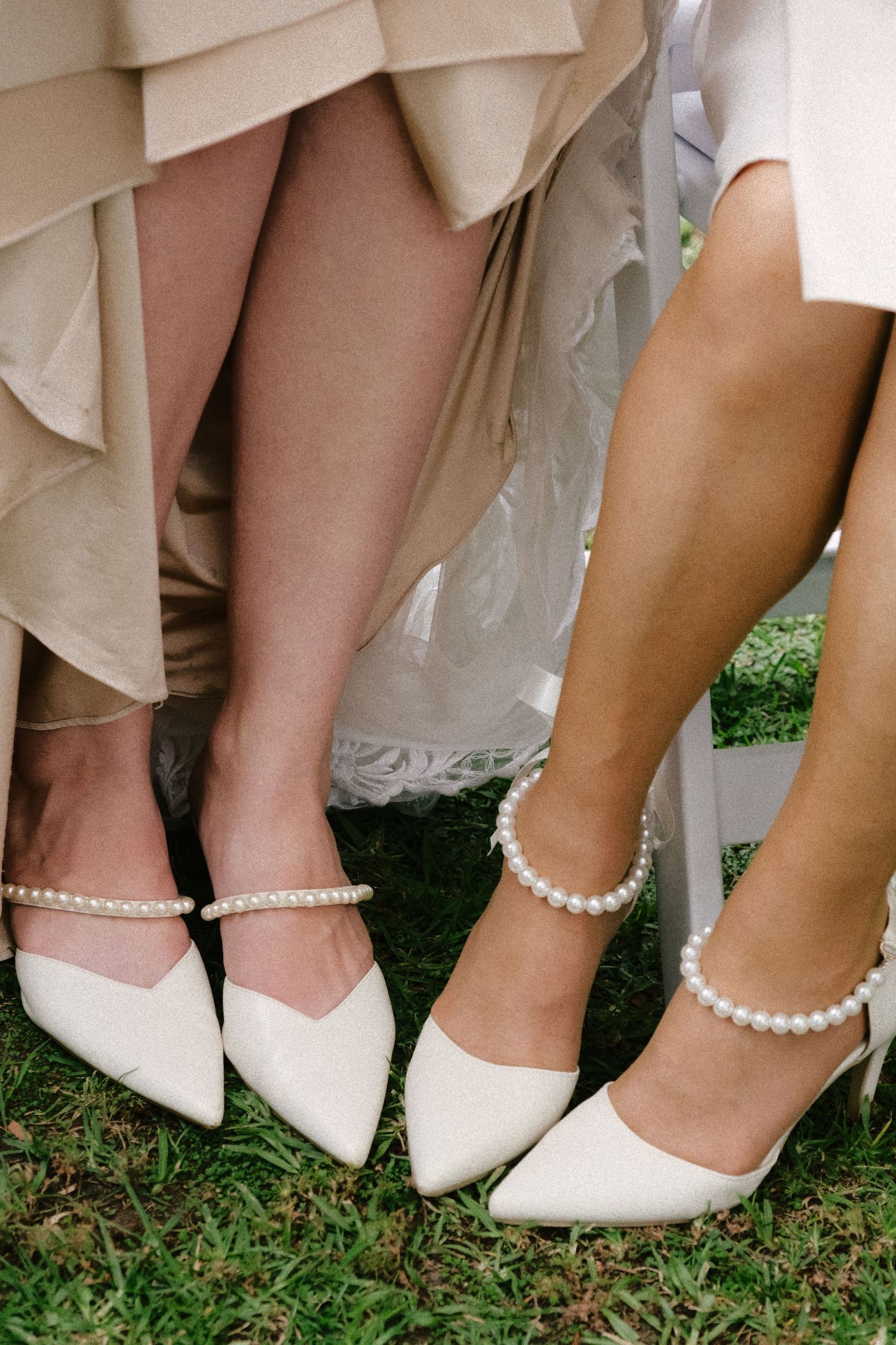 Row of bridesmaids showing off white heels and pearl ankle bracelets while standing on grass.