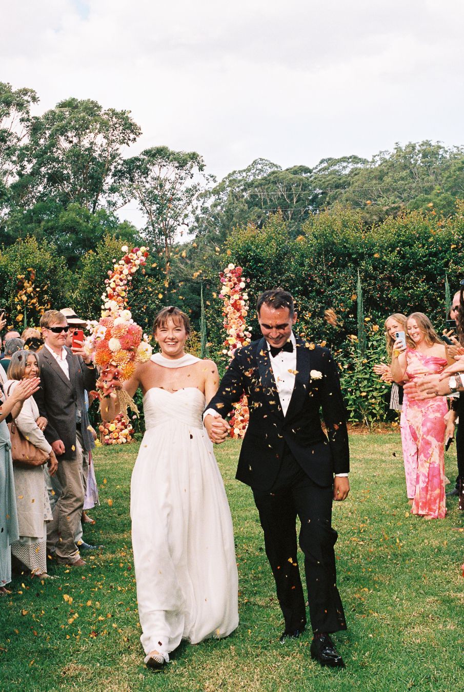 Bride and groom walk down outdoor aisle as guests toss confetti at garden wedding ceremony.