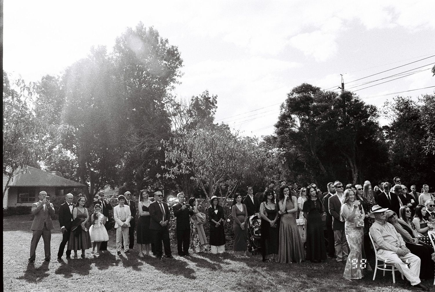 Black and white photo of wedding guests standing in a semi-circle on a lawn beneath tall trees during an outdoor ceremony.