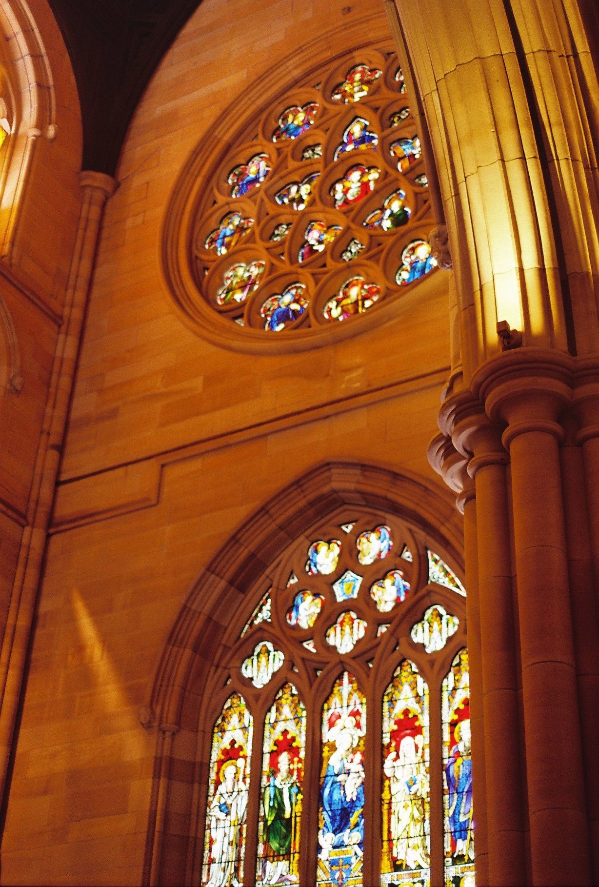 Ornate stained glass windows in a Gothic cathedral illuminate the vaulted ceiling with warm golden light.