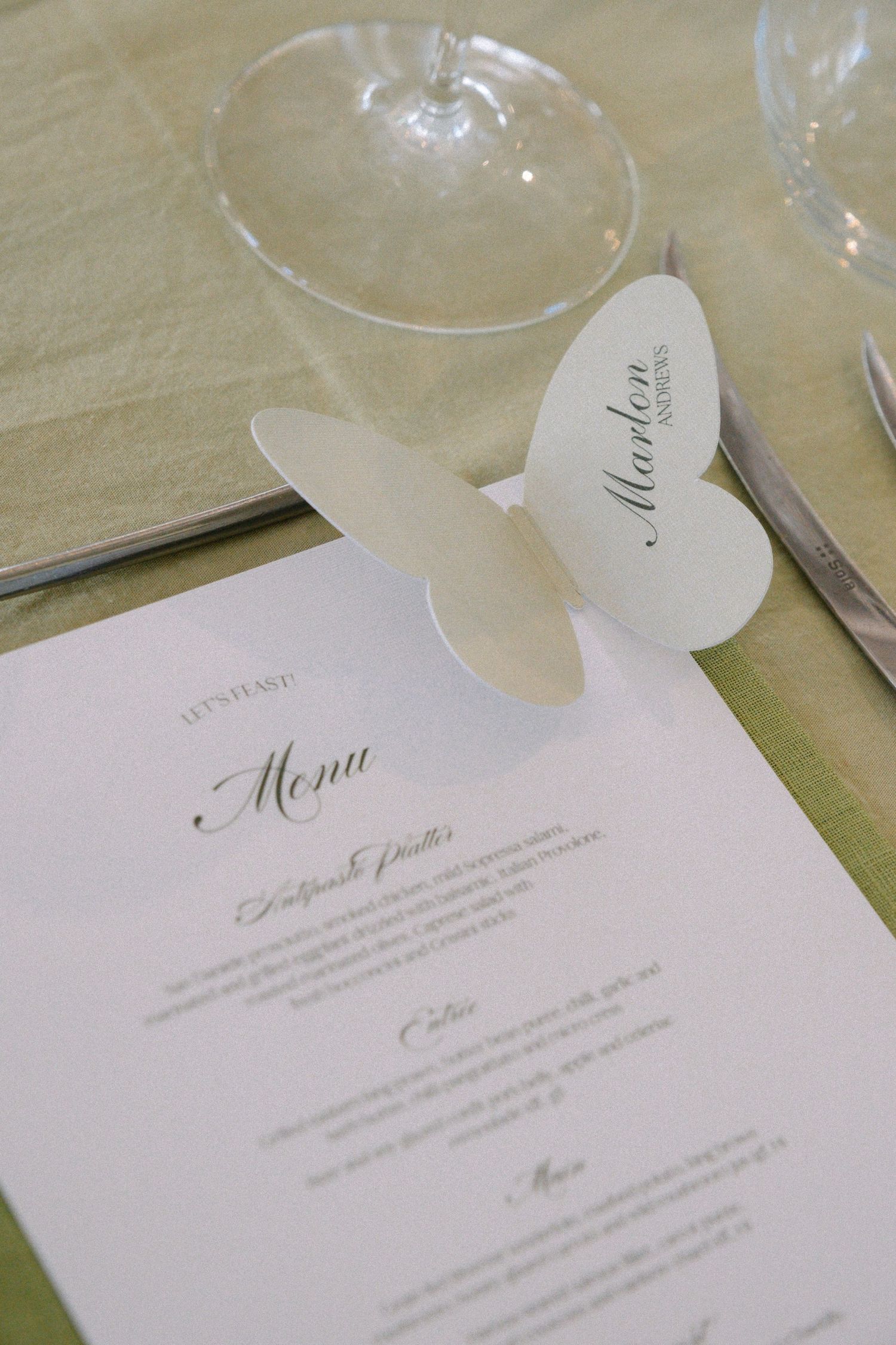 Elegant white wedding menu cards and place cards arranged on a light tablecloth with wine glasses in the background.