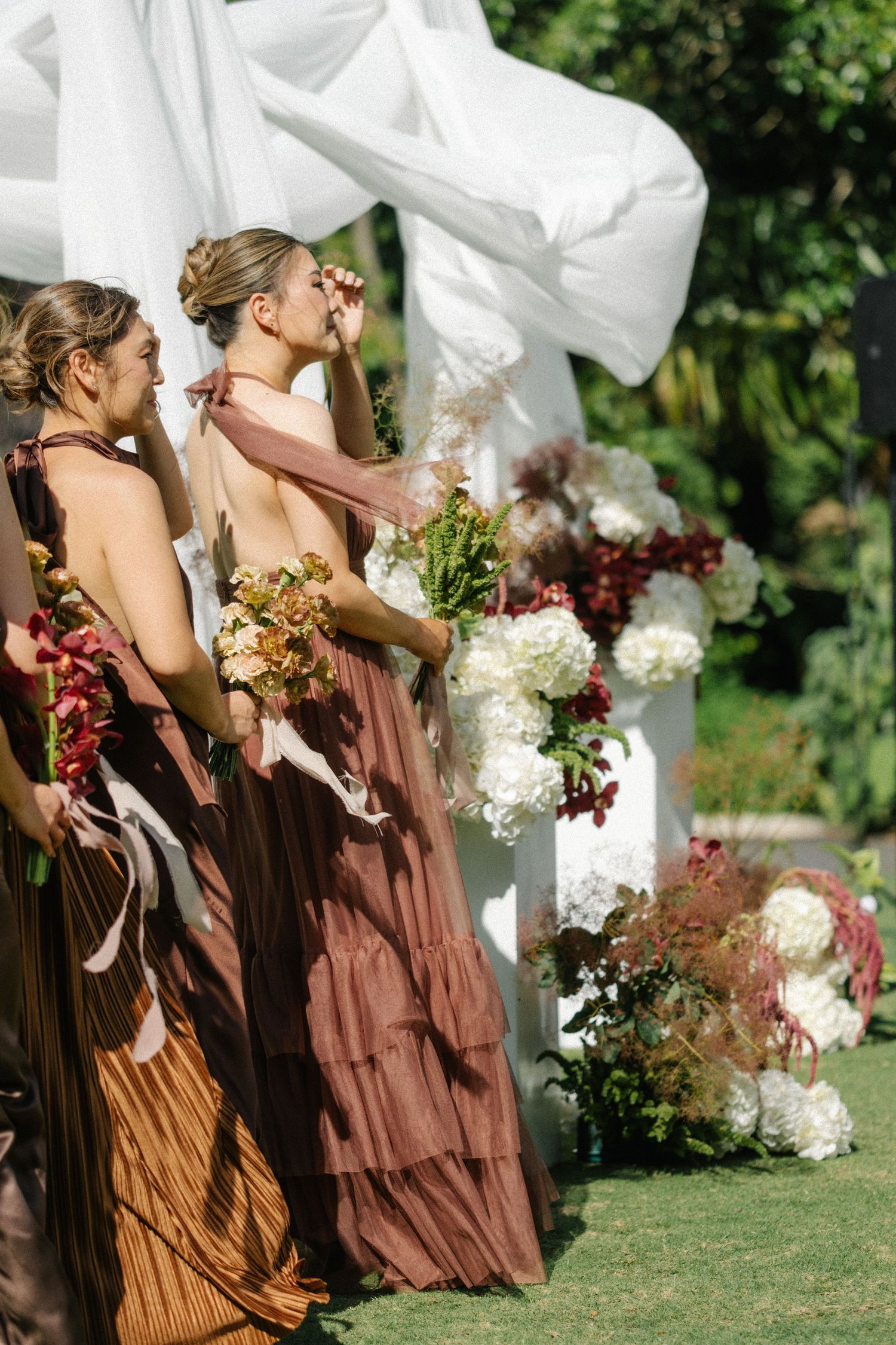 Bridesmaids in flowing brown dresses stand in a line at an outdoor garden wedding ceremony.
