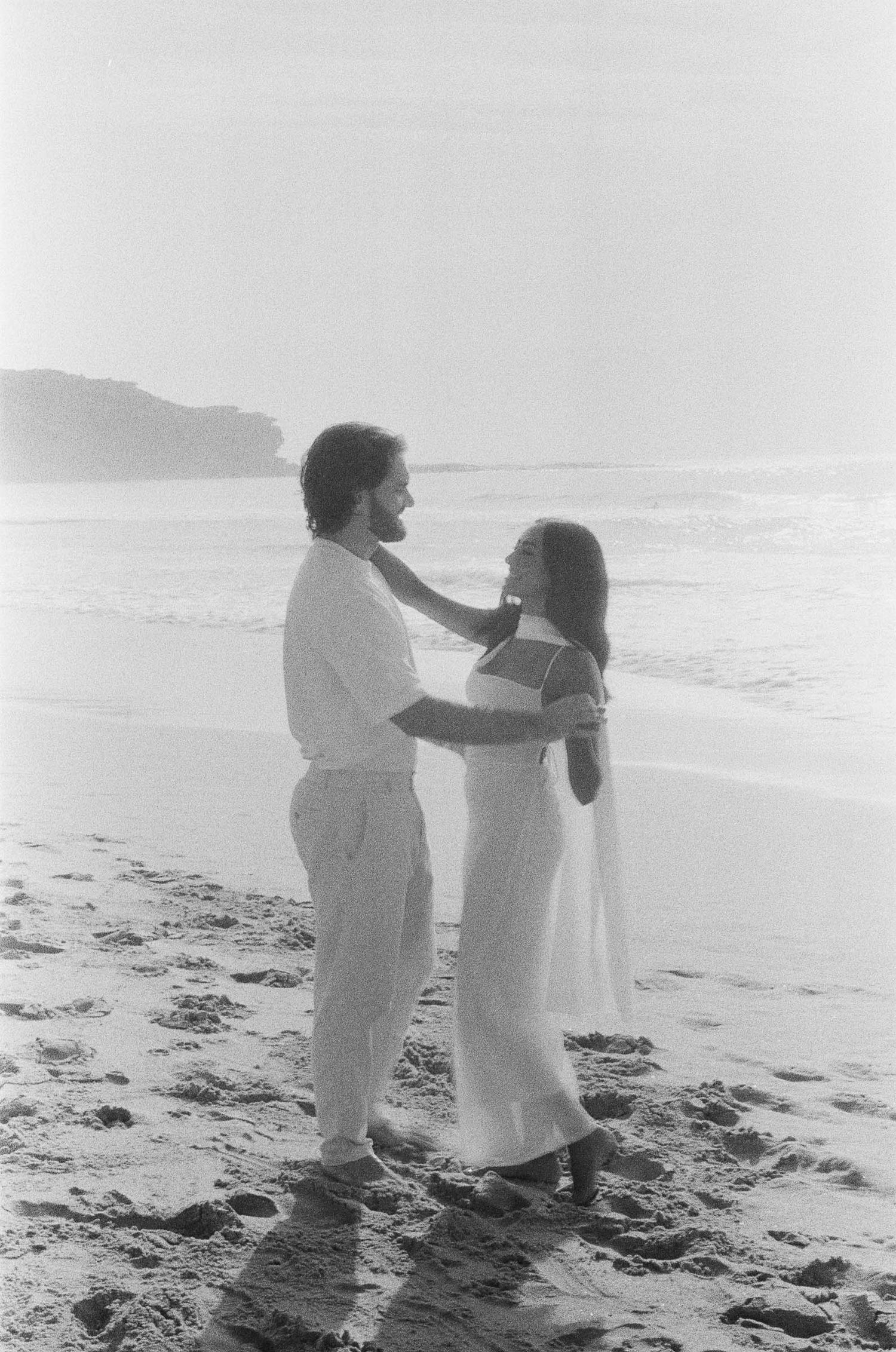 People dressed in white outfits stand together on a misty beach shoreline in black and white photography.