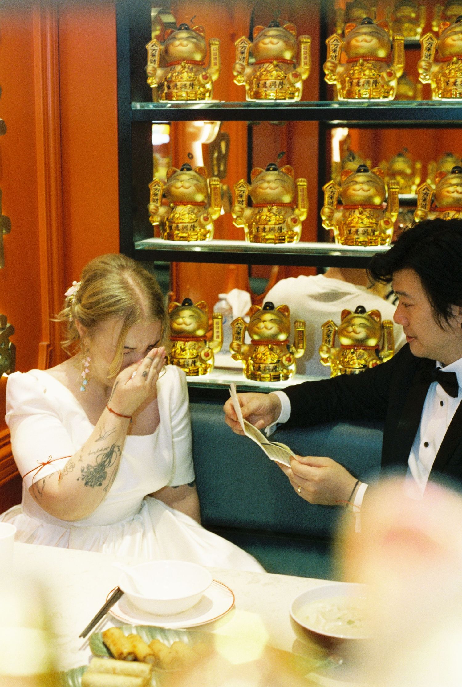 People in formal attire drink from large beer steins at a bar with shelves of golden bottles in the background.