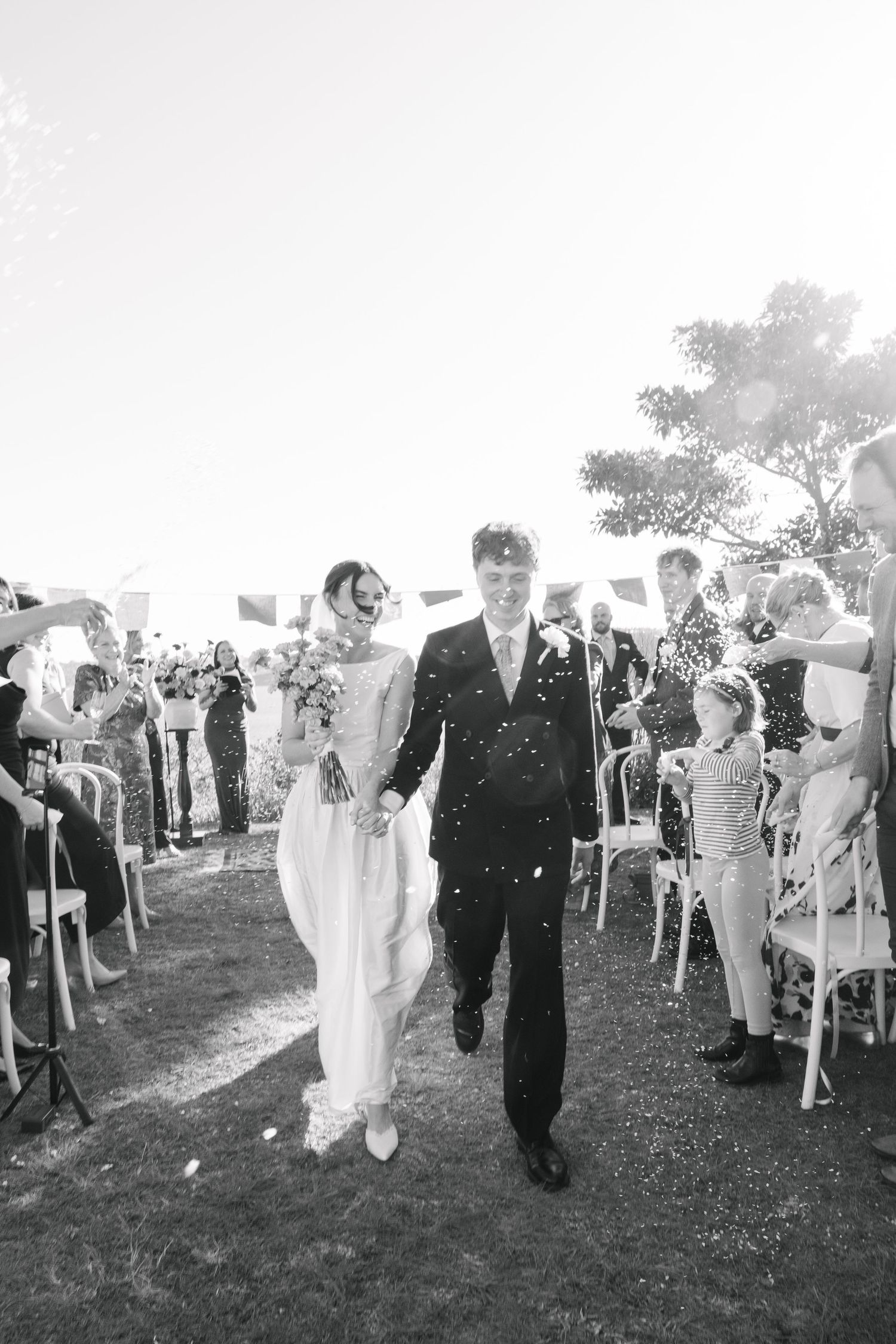Black and white photo of a newlywed couple walking down the aisle as guests throw confetti outdoors.