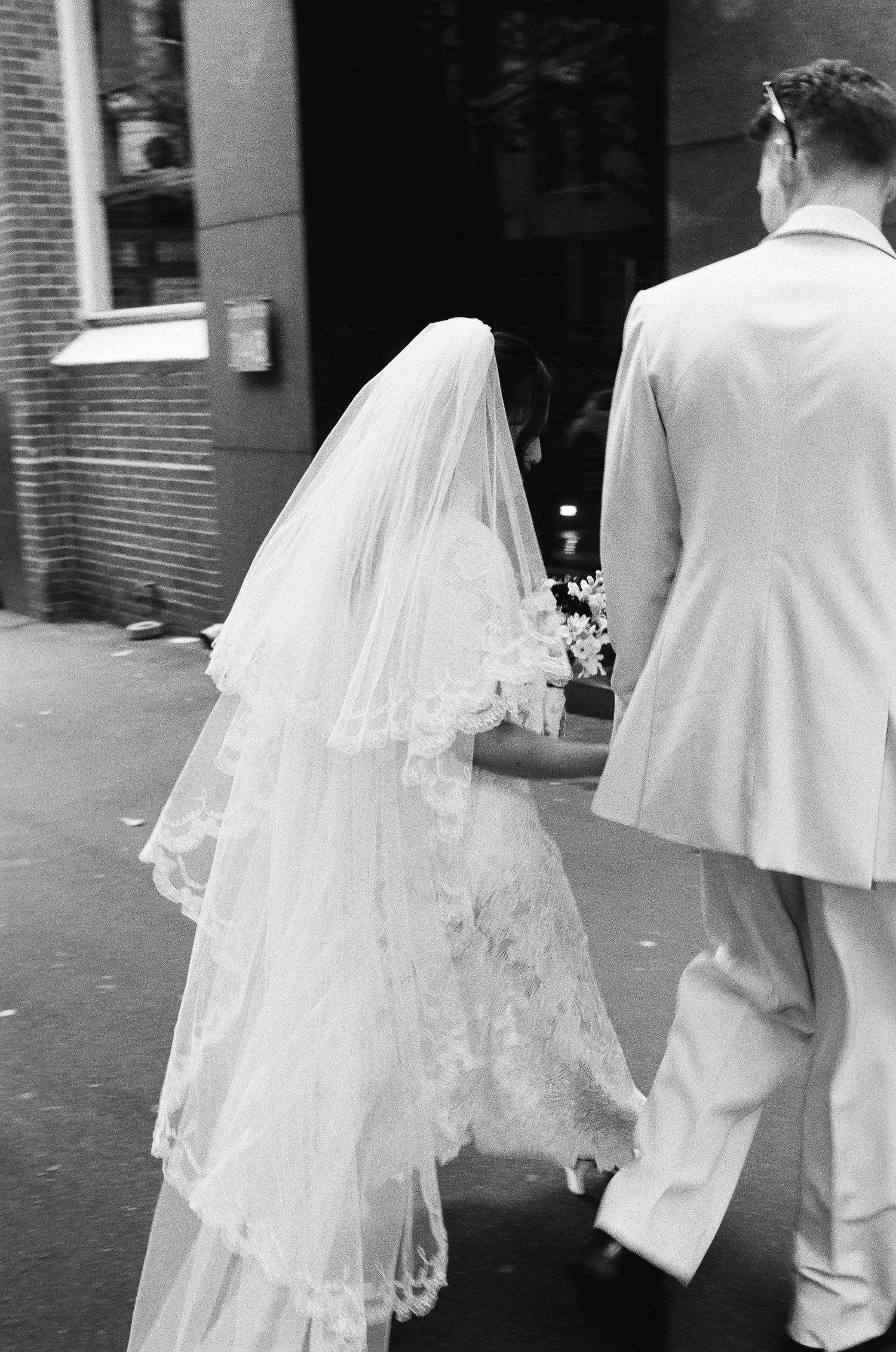 Black and white photo of a bride in a lace veil and gown walking hand in hand with a groom in a light suit past a brick building.