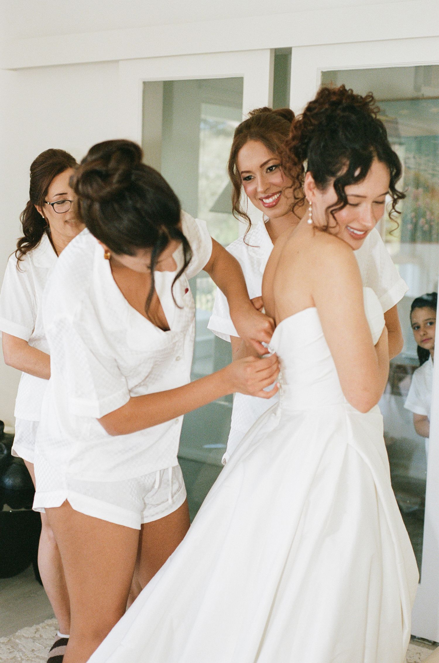 Bridesmaids in matching white pajama sets smile as they help a bride in a strapless white wedding gown get ready before the ceremony.
