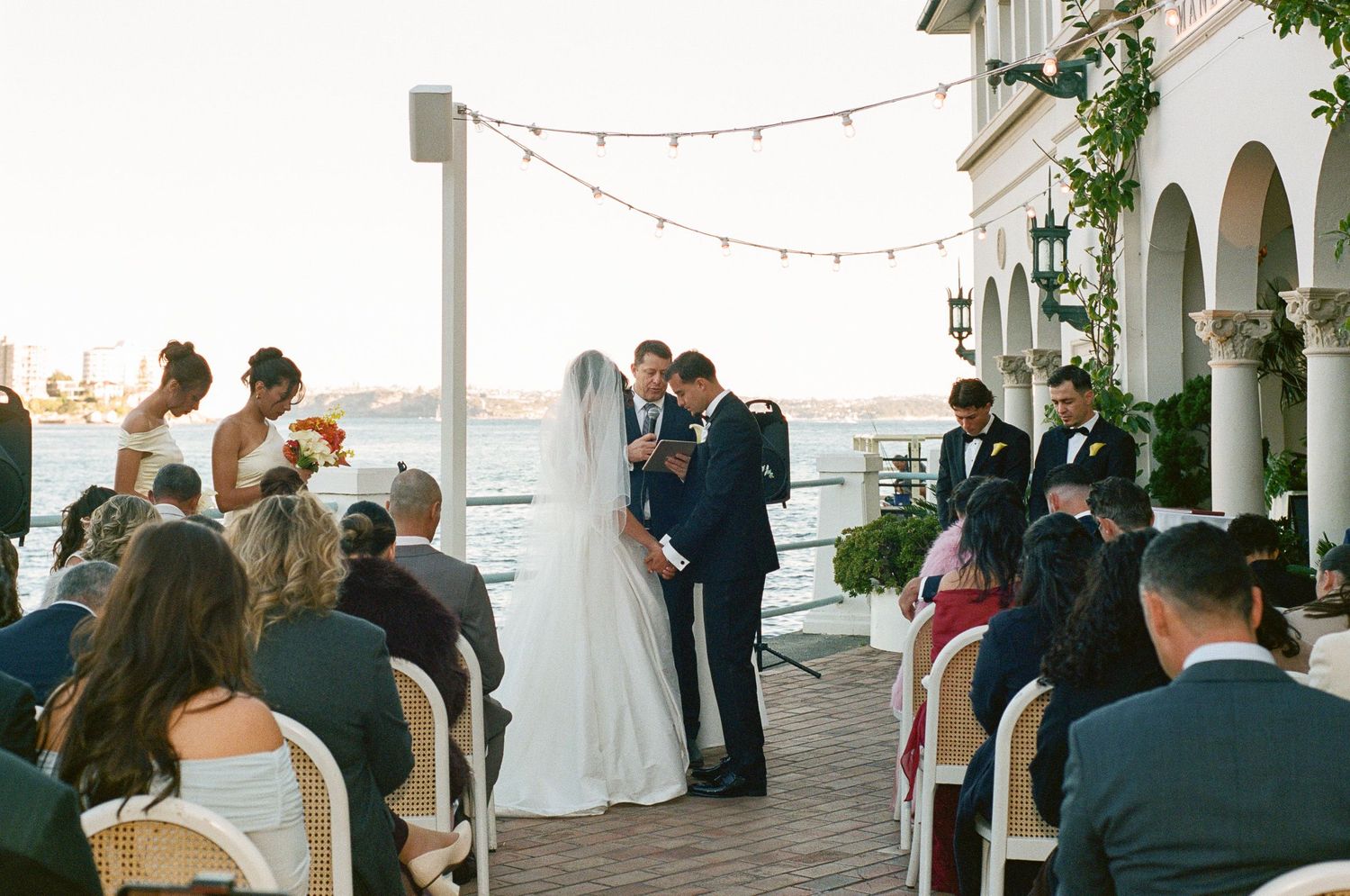 Bride and groom exchange vows during an outdoor waterfront wedding ceremony as seated guests look on beneath string lights.