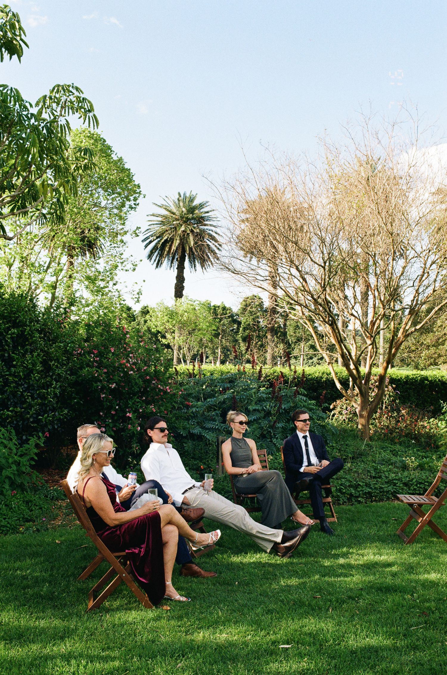 Wedding guests in formal attire sit on wooden folding chairs on a green lawn surrounded by trees and a palm under a blue sky.