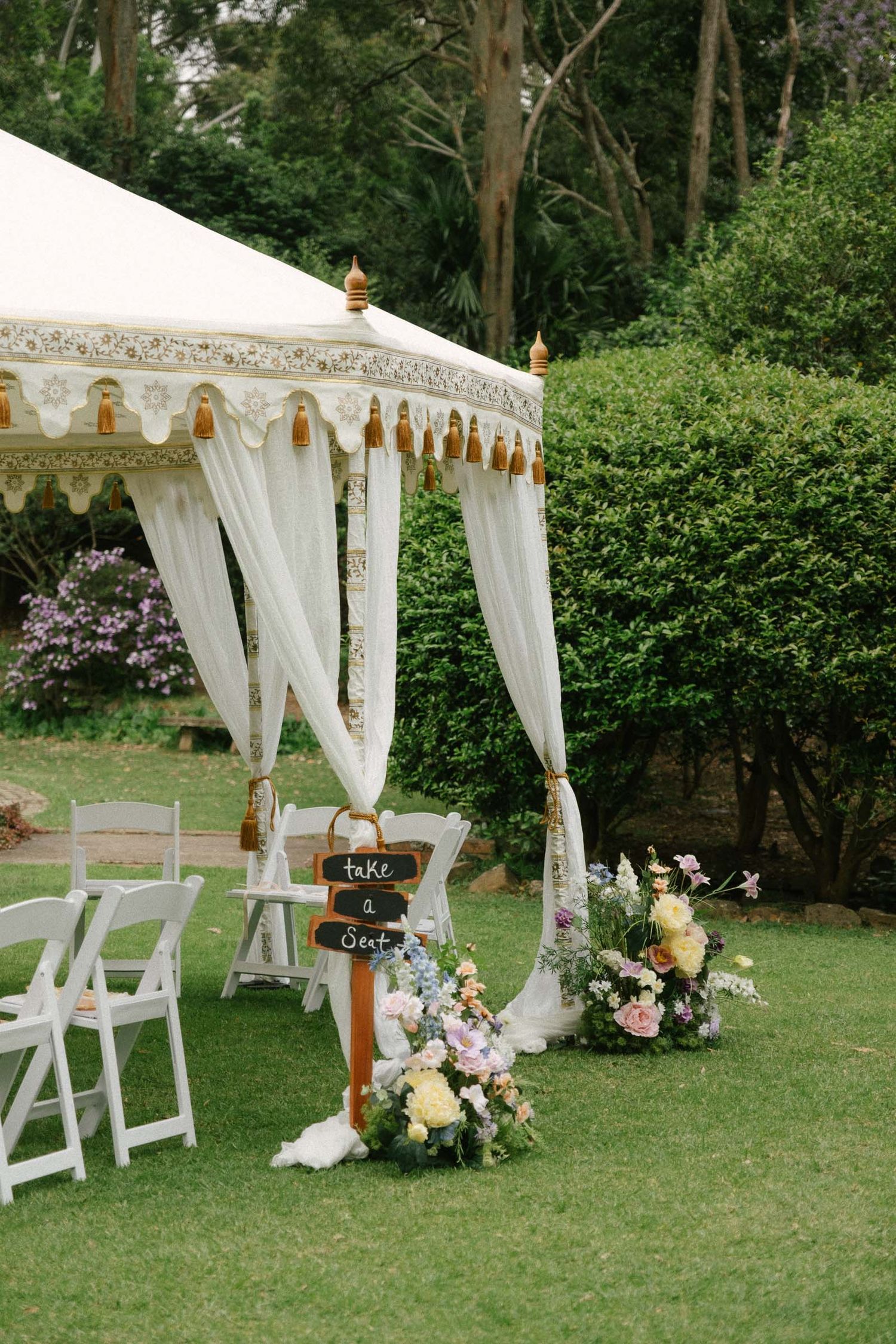 Elegant white wedding ceremony setup with draped canopy, white chairs, and floral arrangements on a green lawn.