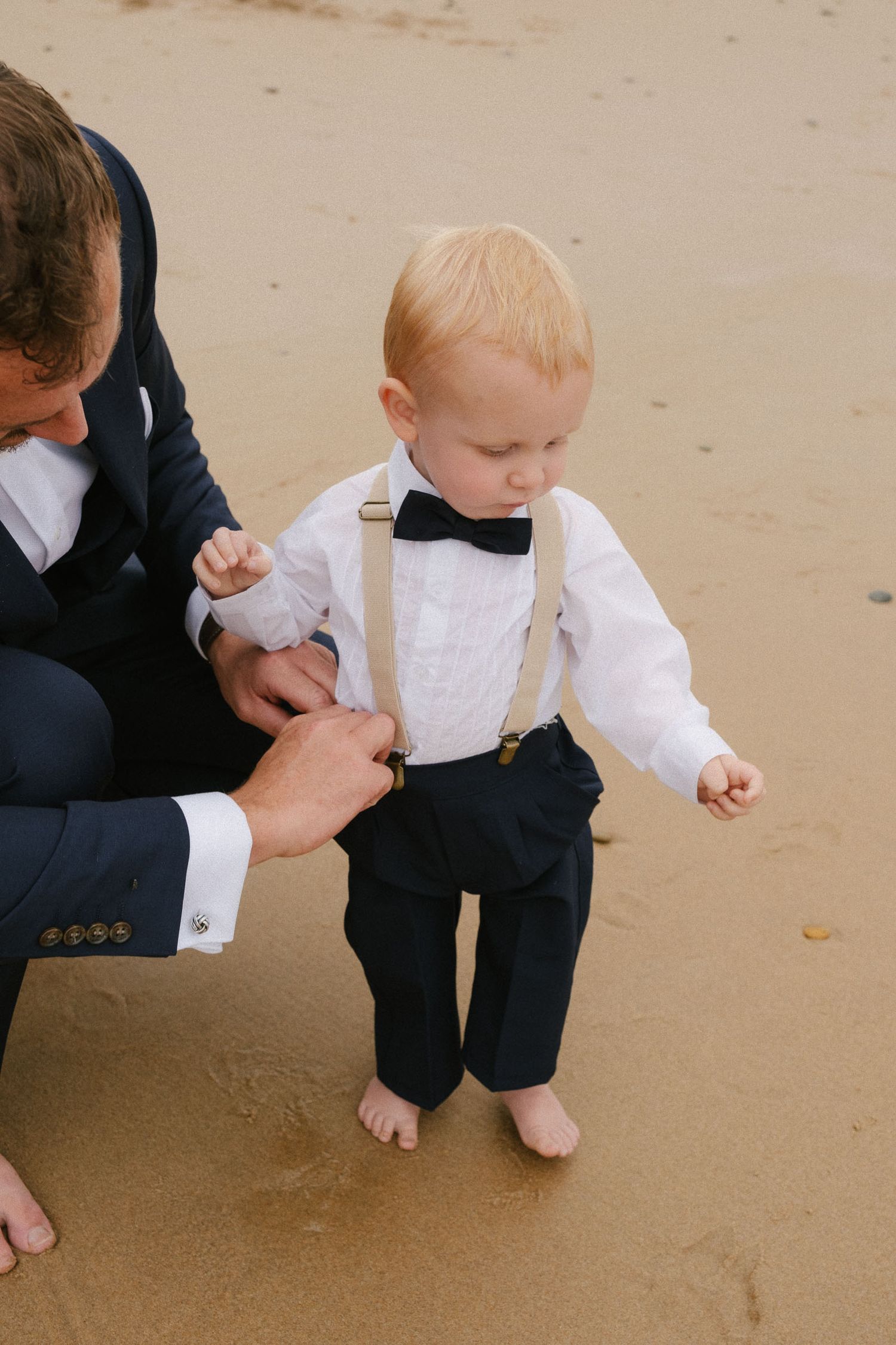 A small child in formal attire with suspenders stands barefoot on the beach while being helped by an adult.