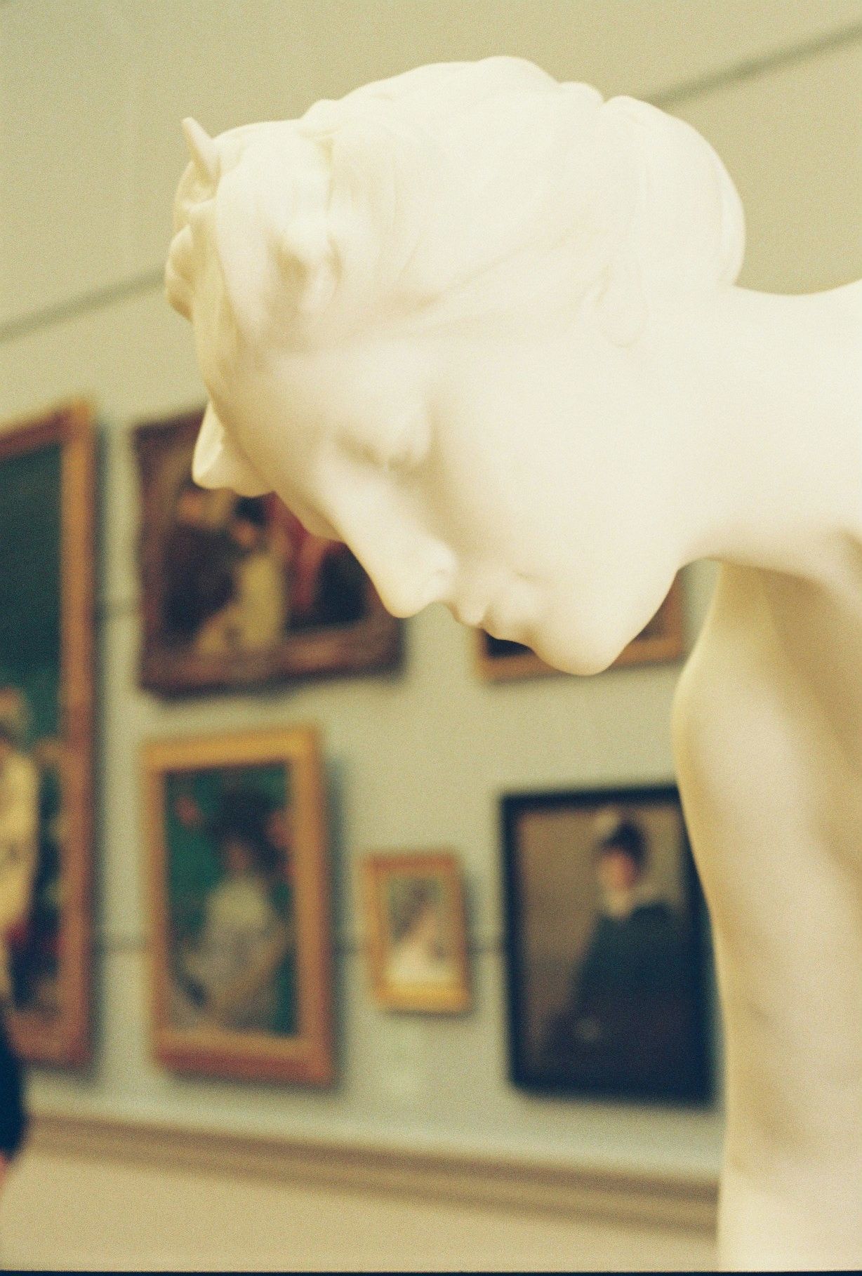 White marble sculpture displayed in an art gallery with framed paintings visible on the wall in soft focus background.