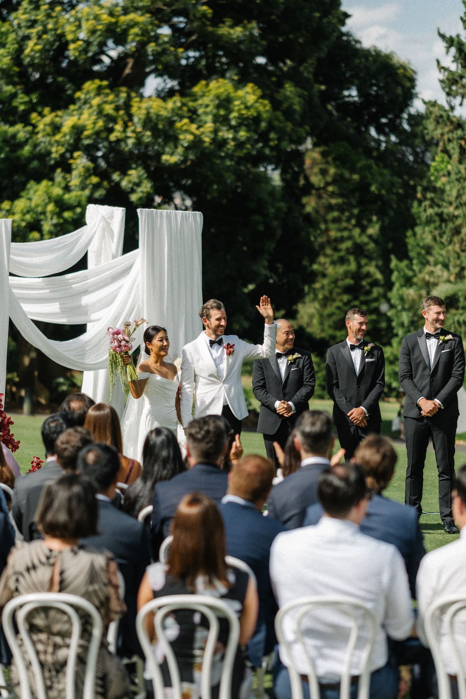 Outdoor wedding ceremony setup with white draped arch and chairs arranged on green lawn in summer garden setting.
