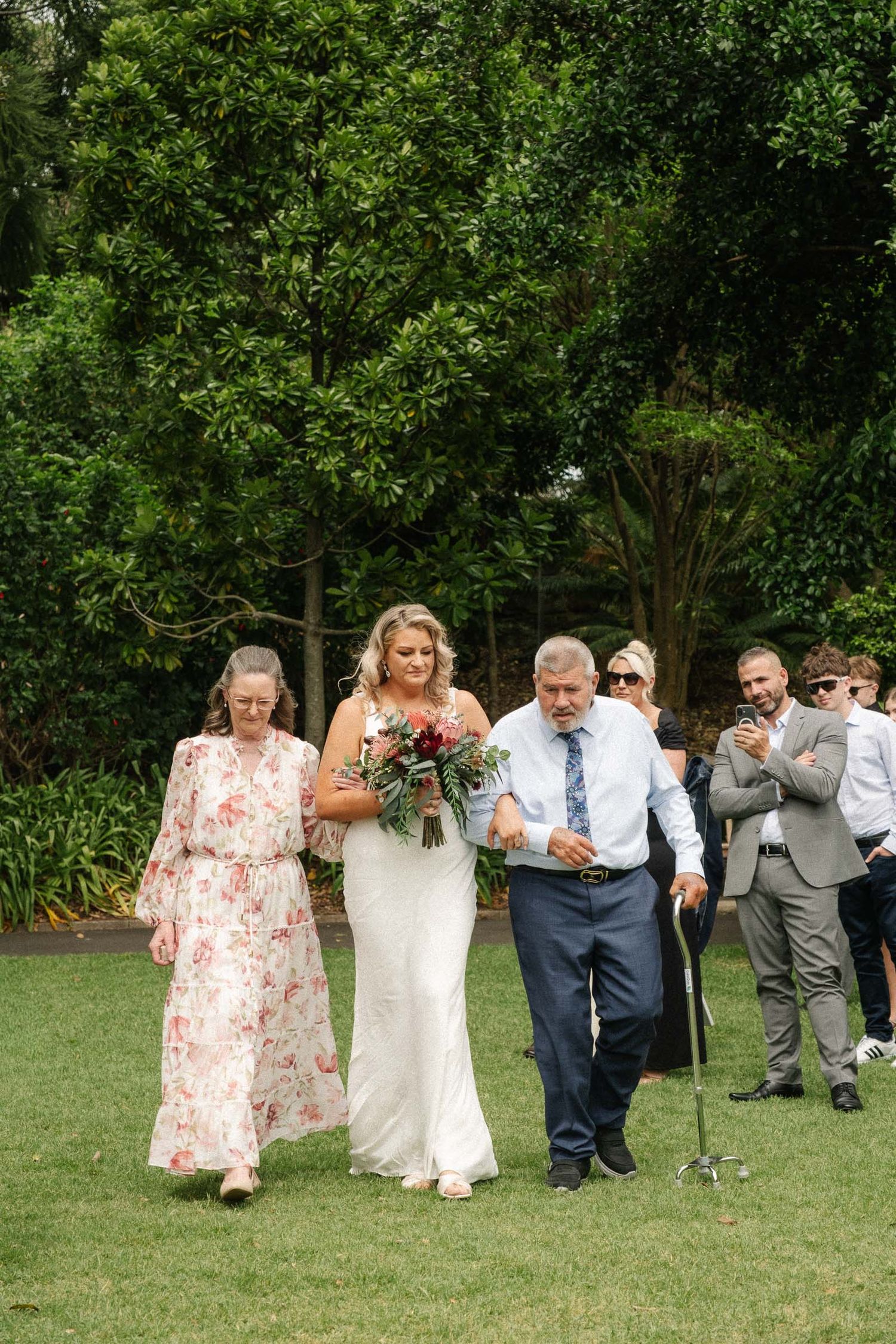 A bride in a white dress walks down a grassy outdoor aisle accompanied by two attendants at a garden wedding ceremony.