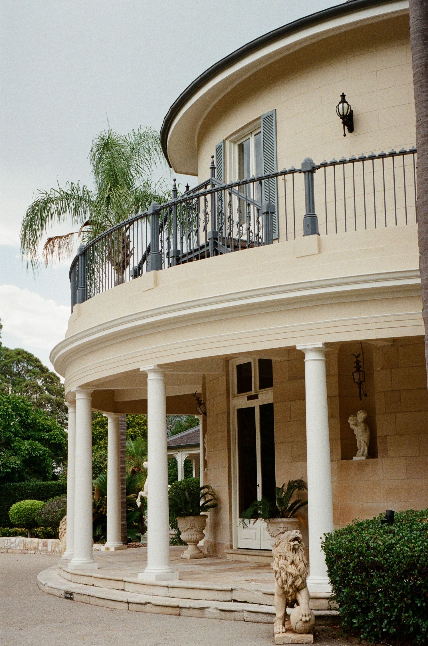 Luxurious Mediterranean-style villa with curved balconies, white columns, and ornate architectural details surrounded by palm trees.