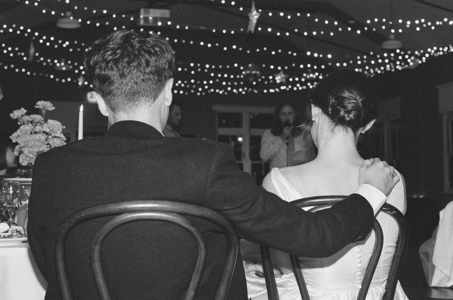 Black and white photo of bride and groom seated at wedding reception table under twinkling string lights.