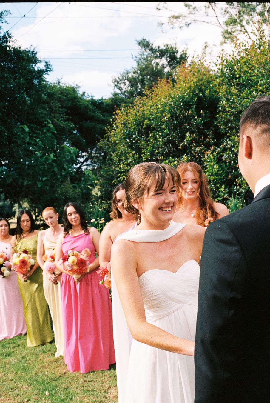 Smiling bride in white strapless gown at outdoor wedding ceremony with colorful bridesmaids in pink and green dresses.