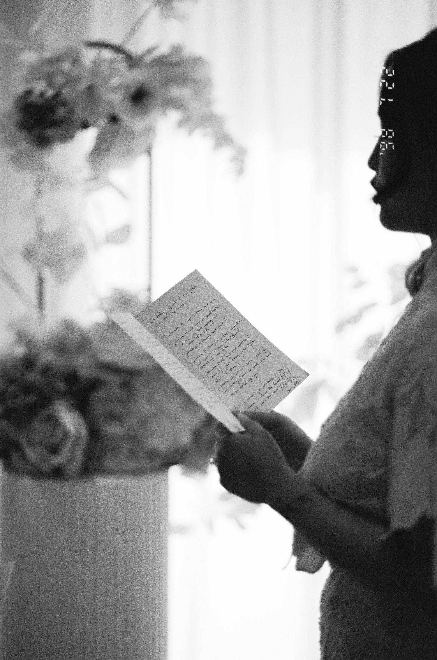 Black and white photo of a woman in profile reading a handwritten letter beside a vase of flowers near a softly lit curtained window.