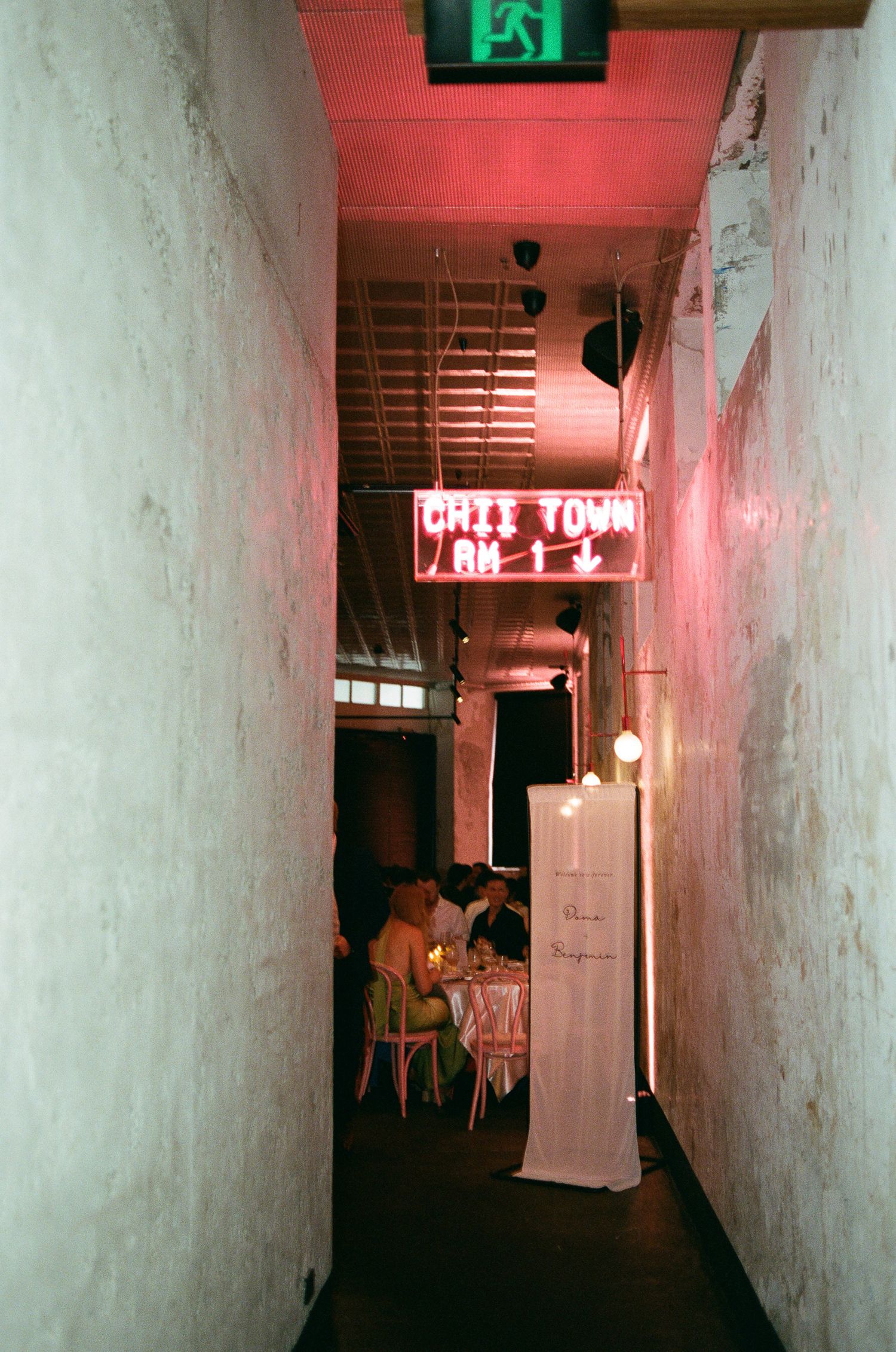 Narrow whitewashed corridor lit by a pink neon Chinatown sign leads to a candlelit dinner table with guests seated in the background.