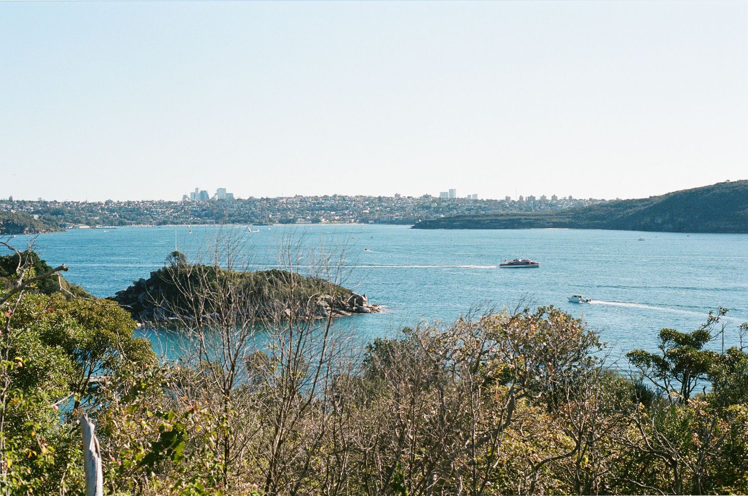Scenic harbor view with a small rocky island, boats on calm blue water, and a distant city skyline beyond wooded green hills.