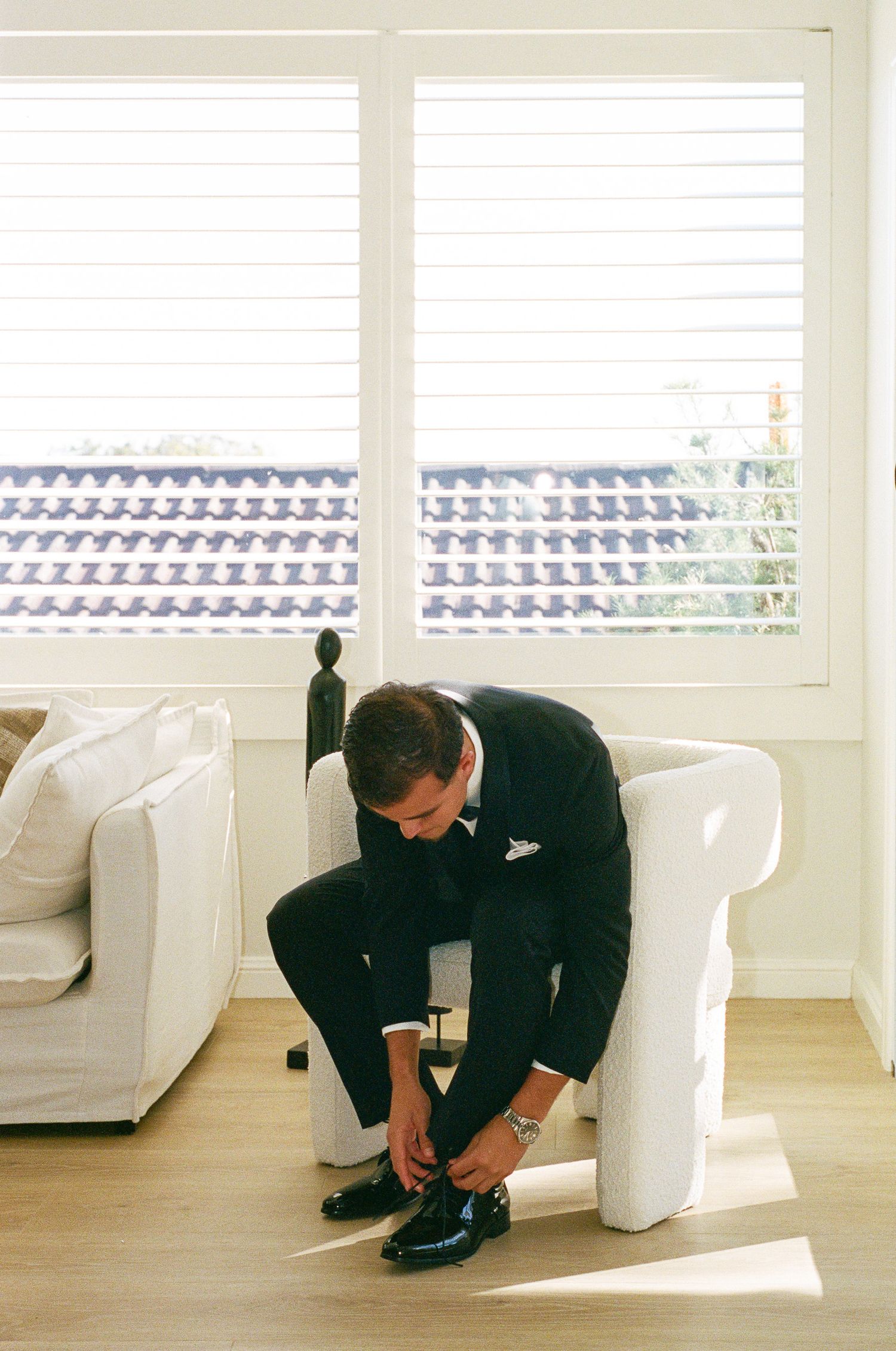 Groom in a black suit sits on a white armchair tying his dress shoes in a bright room with white shutters and tiled rooftops outside.