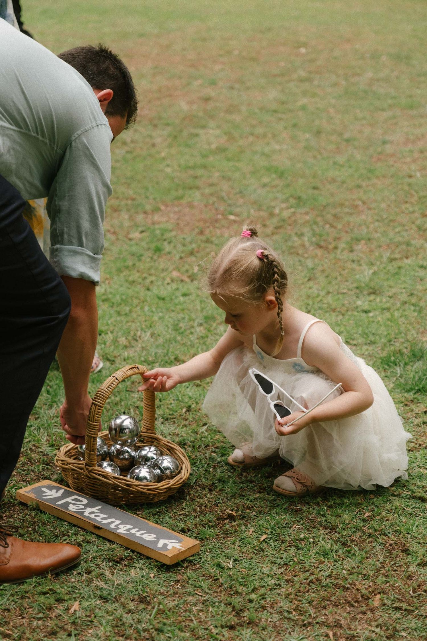 A young flower girl in white dress crouches down to look at decorative items in a basket on the grass.